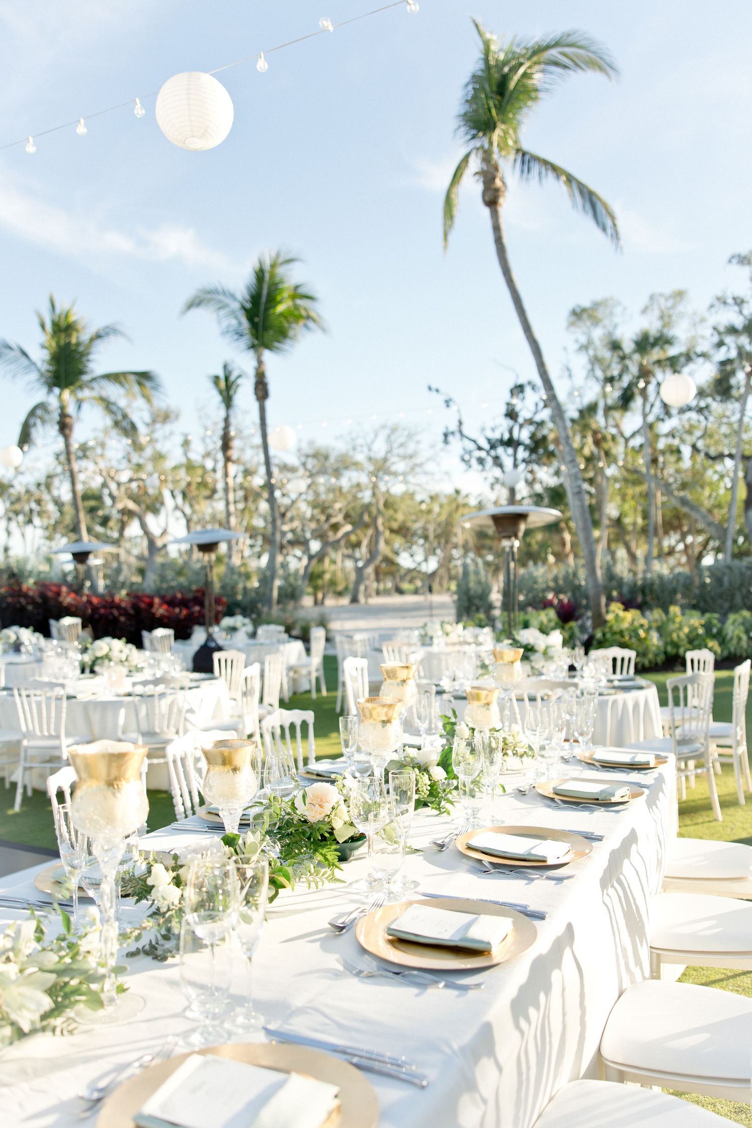Elegant outdoor reception tables with white chairs and tropical palm trees.