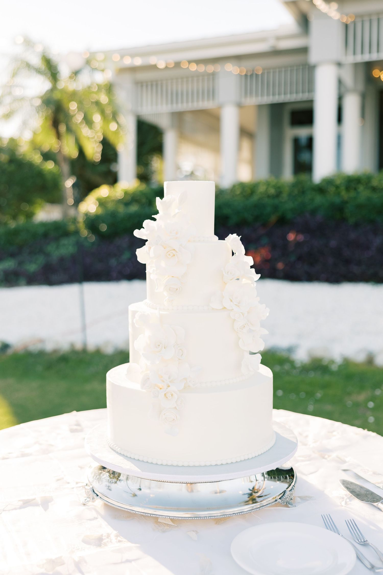 An elegant four-tier white wedding cake decorated with delicate floral details stands outdoors.
