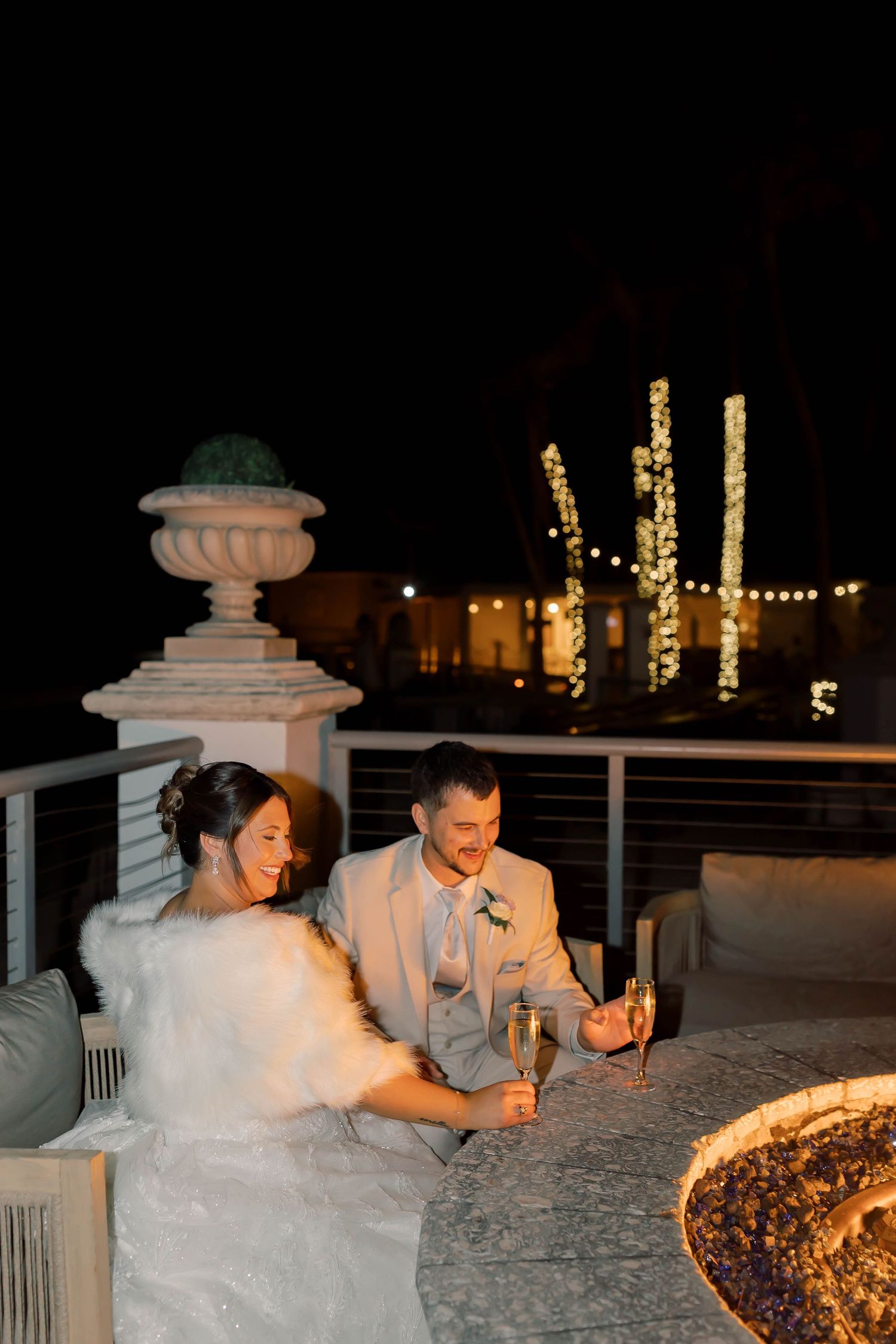 Bride and groom toast champagne by a fire pit on a romantic outdoor terrace at night, with string lights glowing in the background.