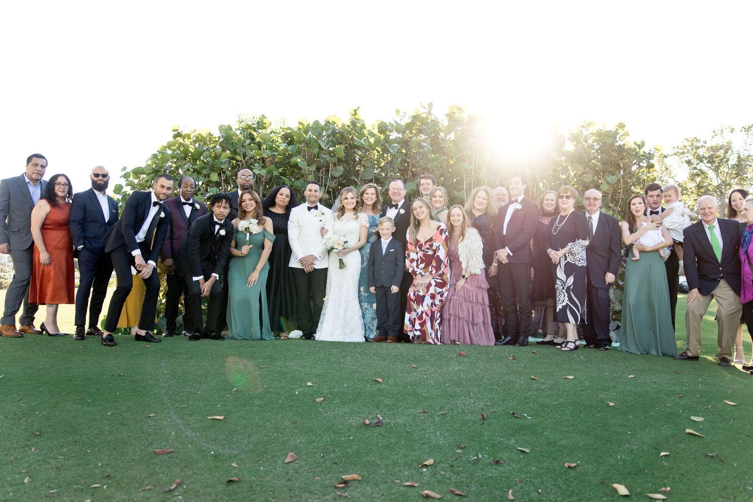 Large wedding party and guests pose for a panoramic group photo on a grassy lawn.