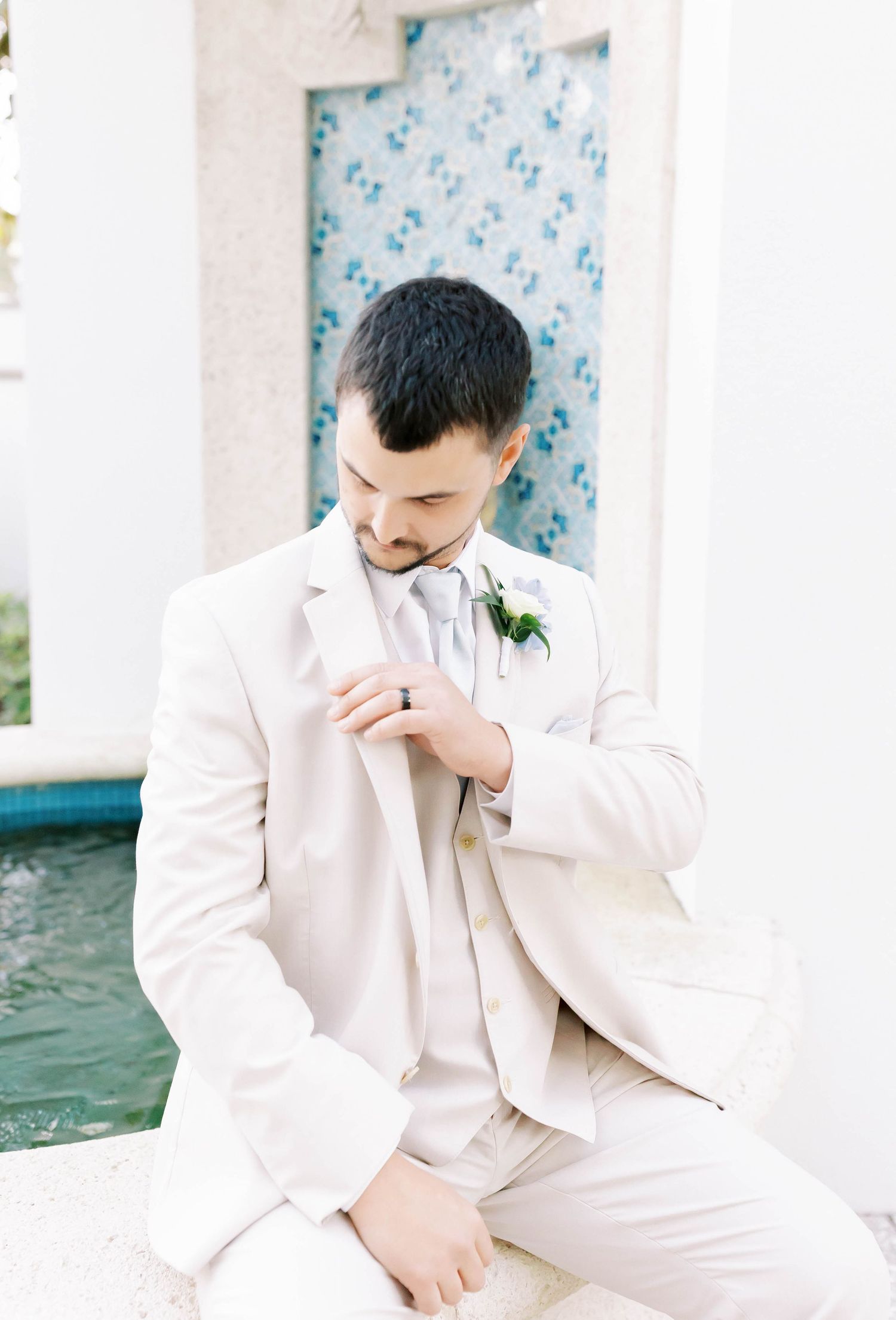 Dapper groom in cream suit adjusts jacket near decorative tiled fountain backdrop.