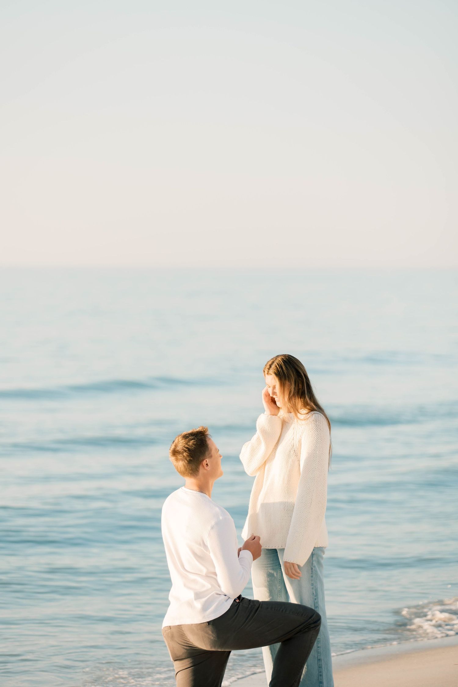 Two people share a tender moment while sitting on a beach with gentle waves in the background.
