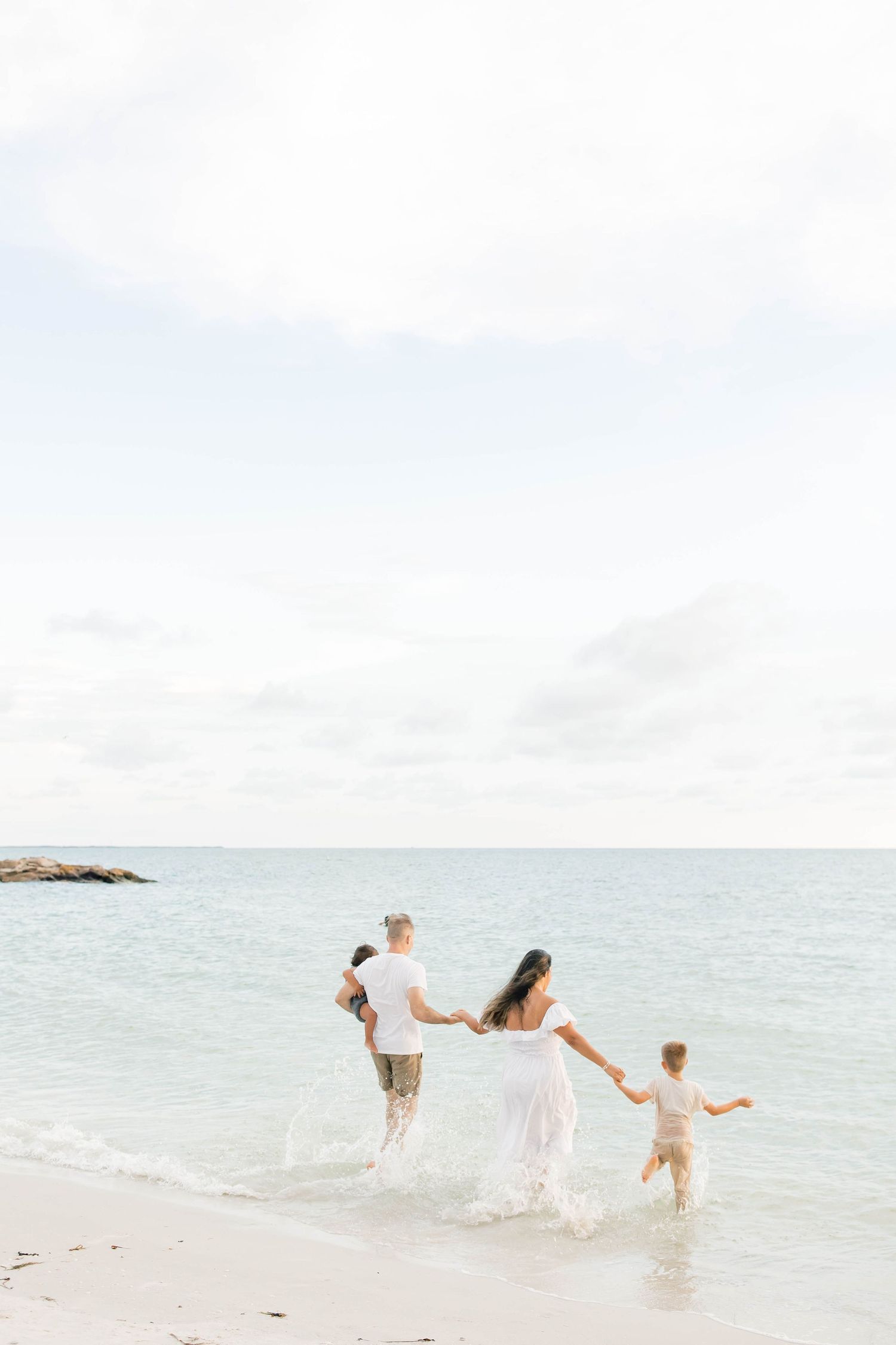 A family walks hand in hand along the shoreline in white outfits during a beach photoshoot.