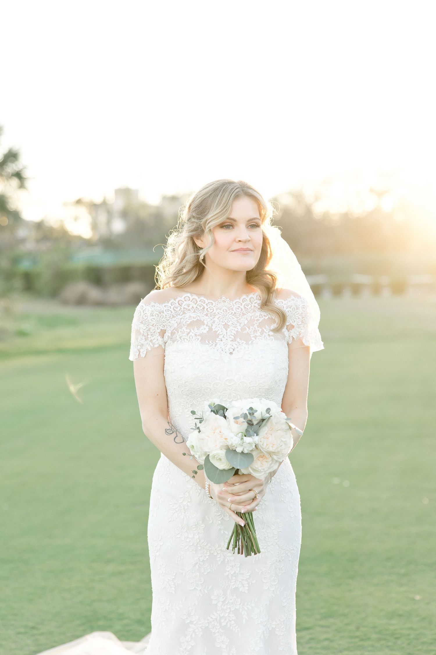 Bride in lace dress holding white floral bouquet against sunset on golf course.