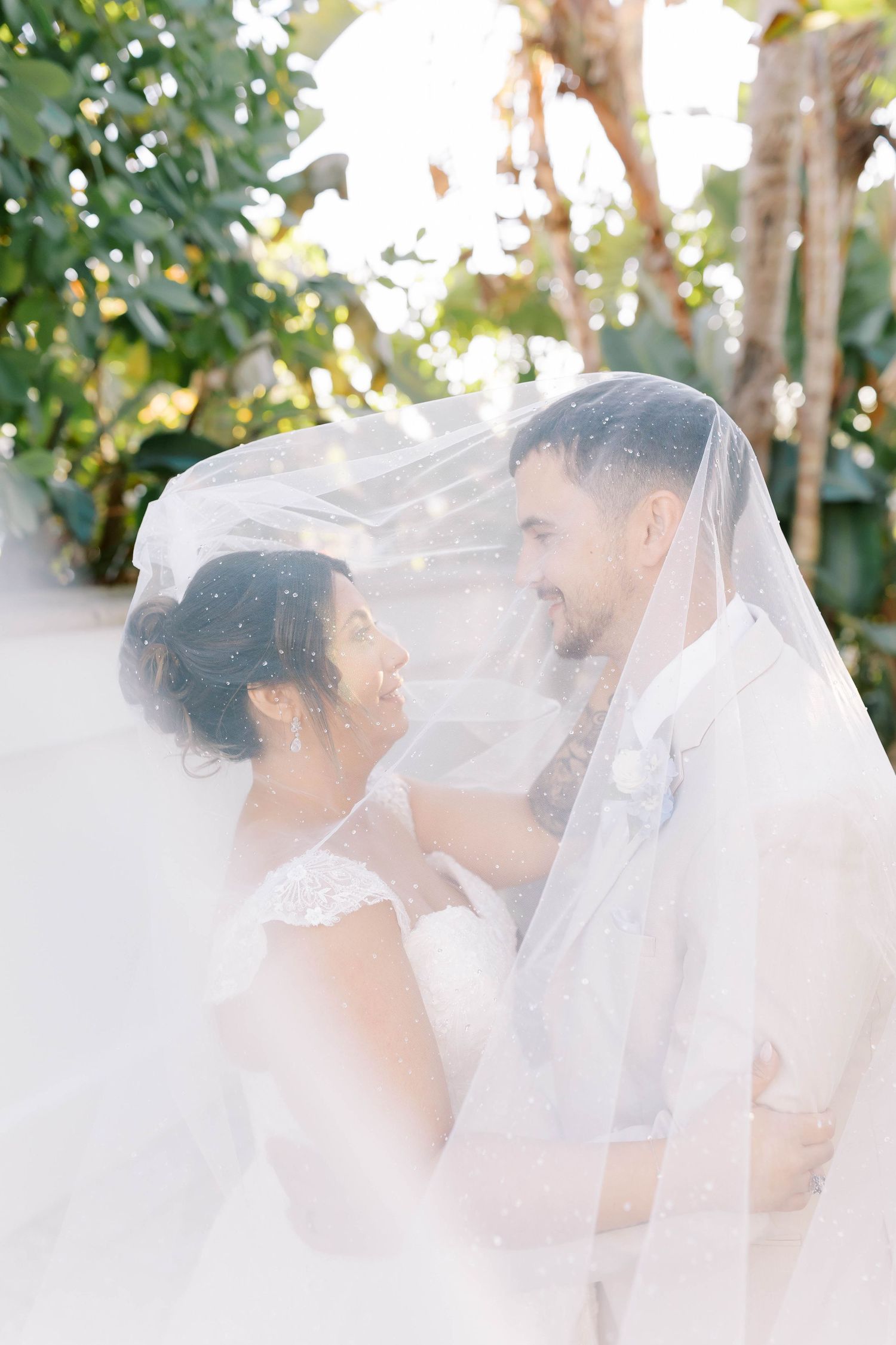 Bride and groom smiling under a sheer wedding veil surrounded by lush tropical greenery.
