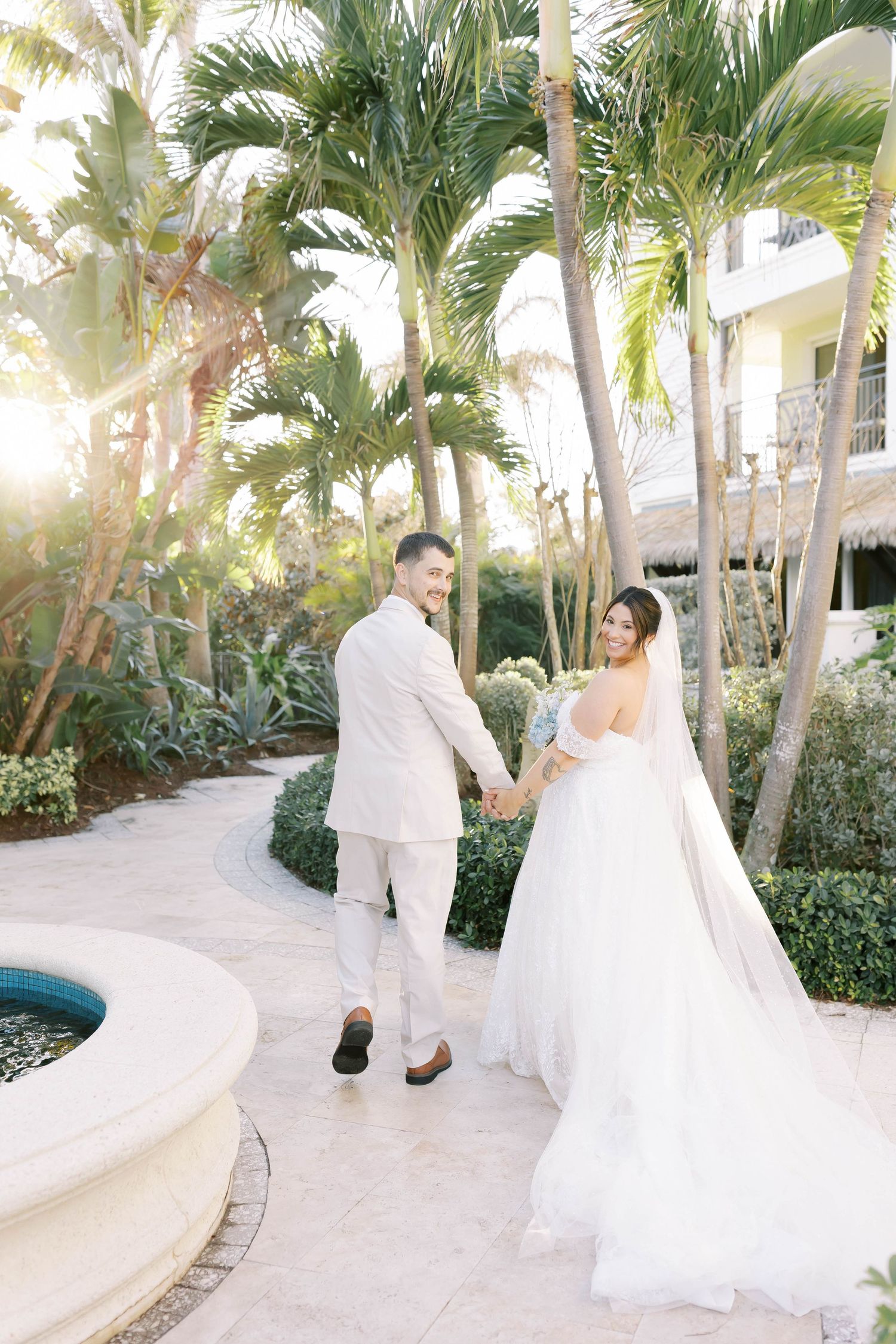 Bride and groom hold hands walking along a tropical resort pathway surrounded by palm trees in golden sunlight.