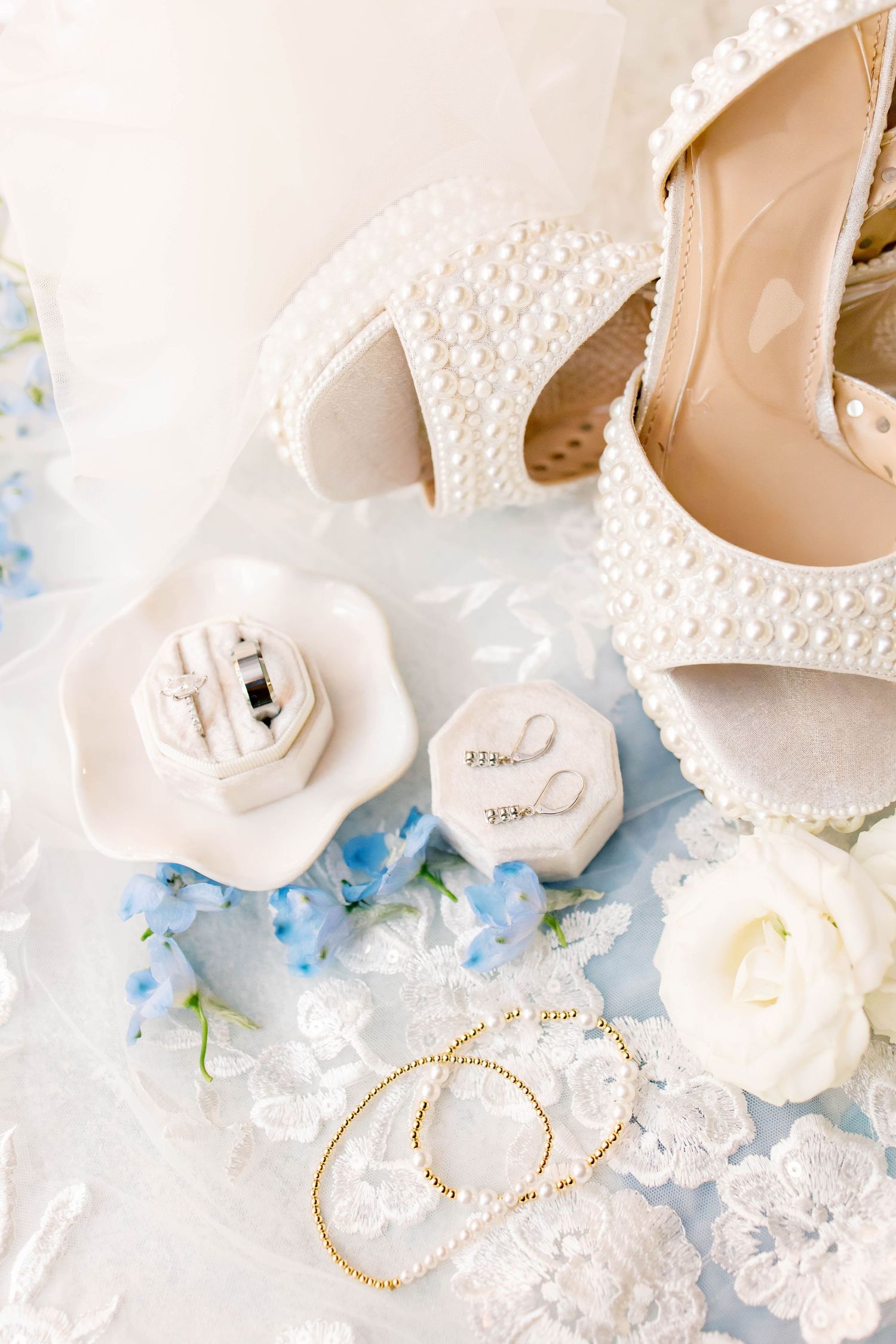 Pearl-embellished bridal heels displayed with ring boxes, gold hoop earrings, white rose, and blue flowers on lace fabric.
