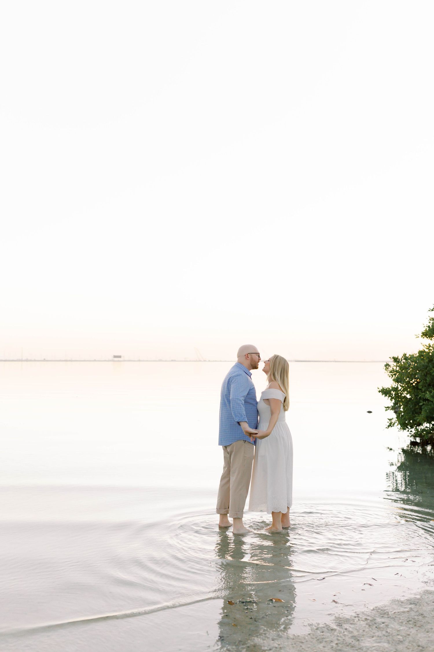 A couple stands together in shallow water at the edge of a serene beach during golden hour.