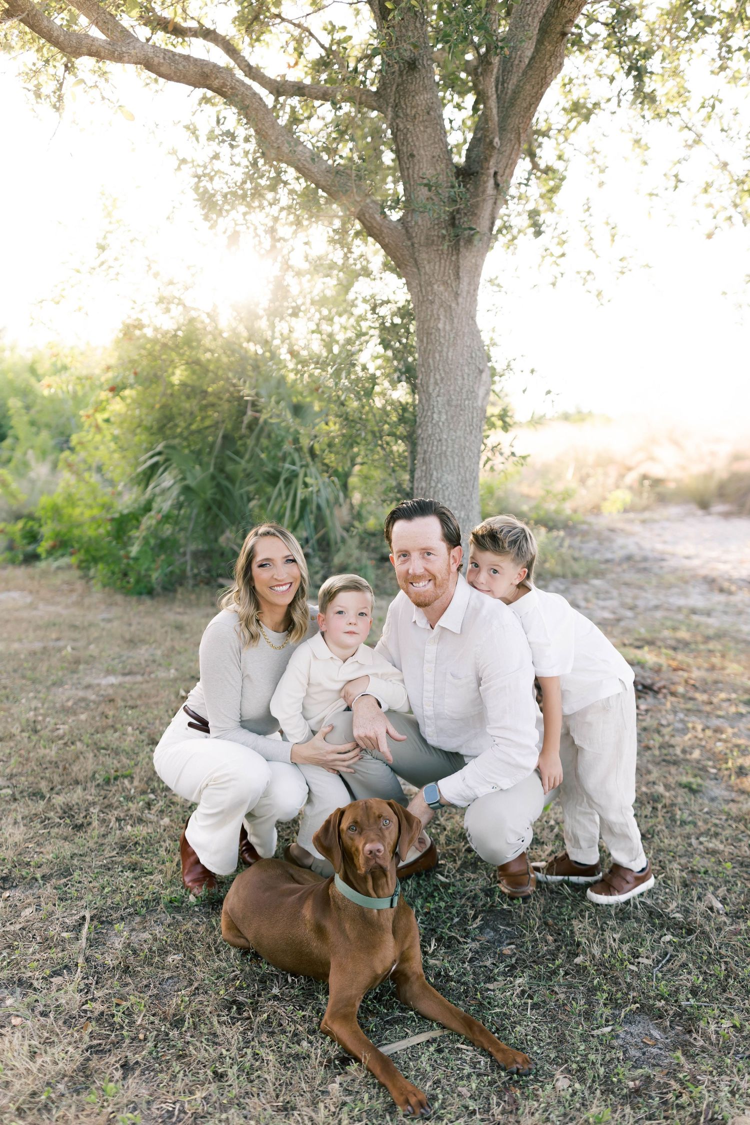 Family dressed in white sits under a tree with their brown dog during a photo session.