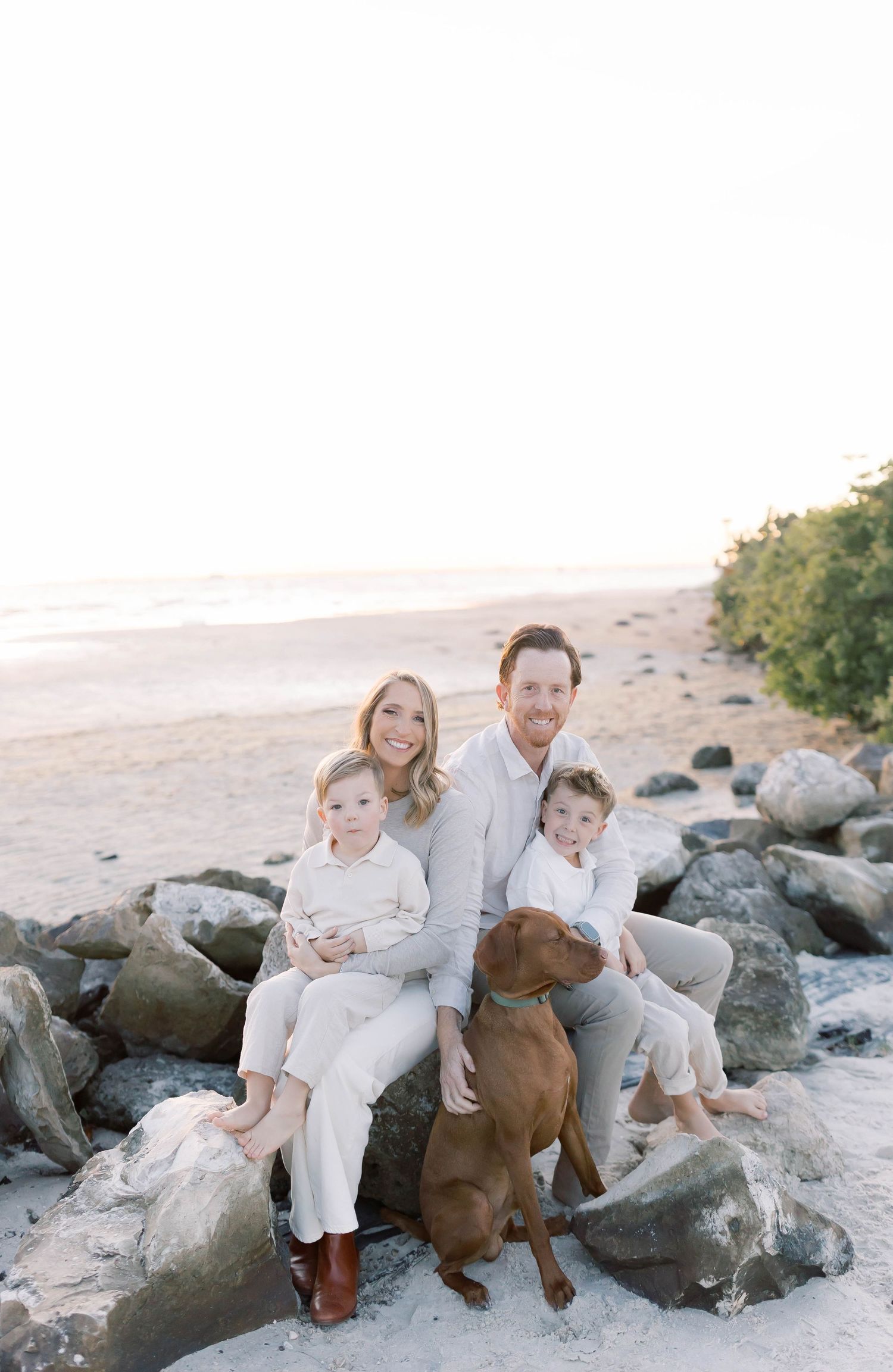 Family and their brown dog pose together on rocky beach in white and neutral clothing.