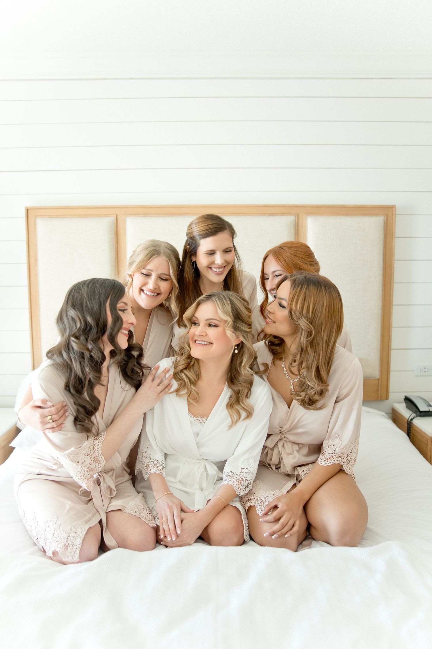 A group of women in silk robes share a joyful moment together on a white bed.