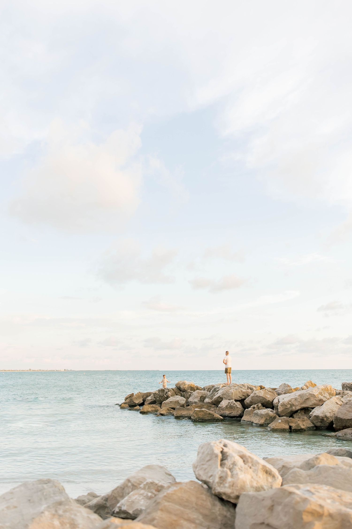 Rocky jetty extending into calm blue waters under a bright cloudy sky.