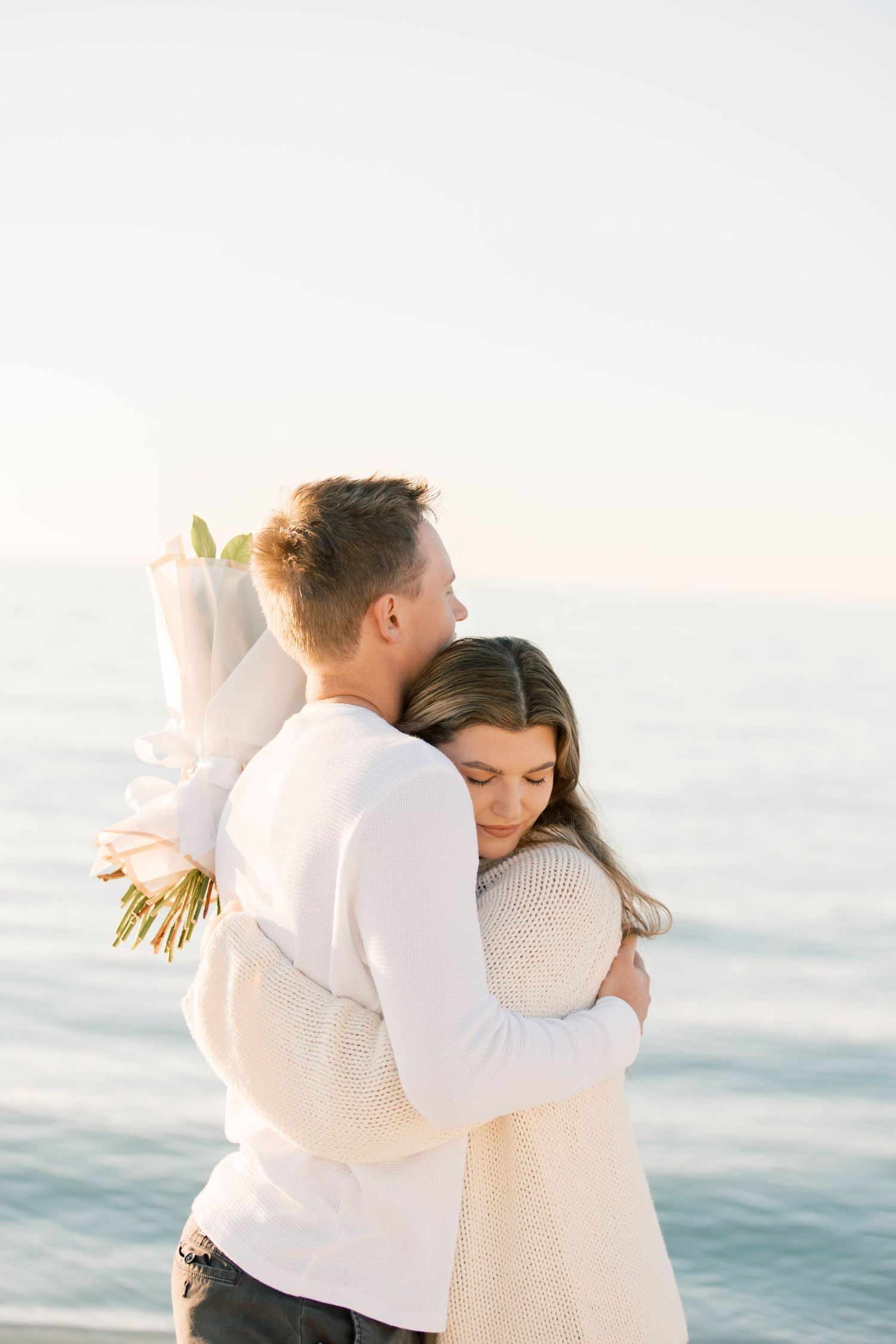 A romantic beach embrace with a couple in light colored clothing holding a bouquet.