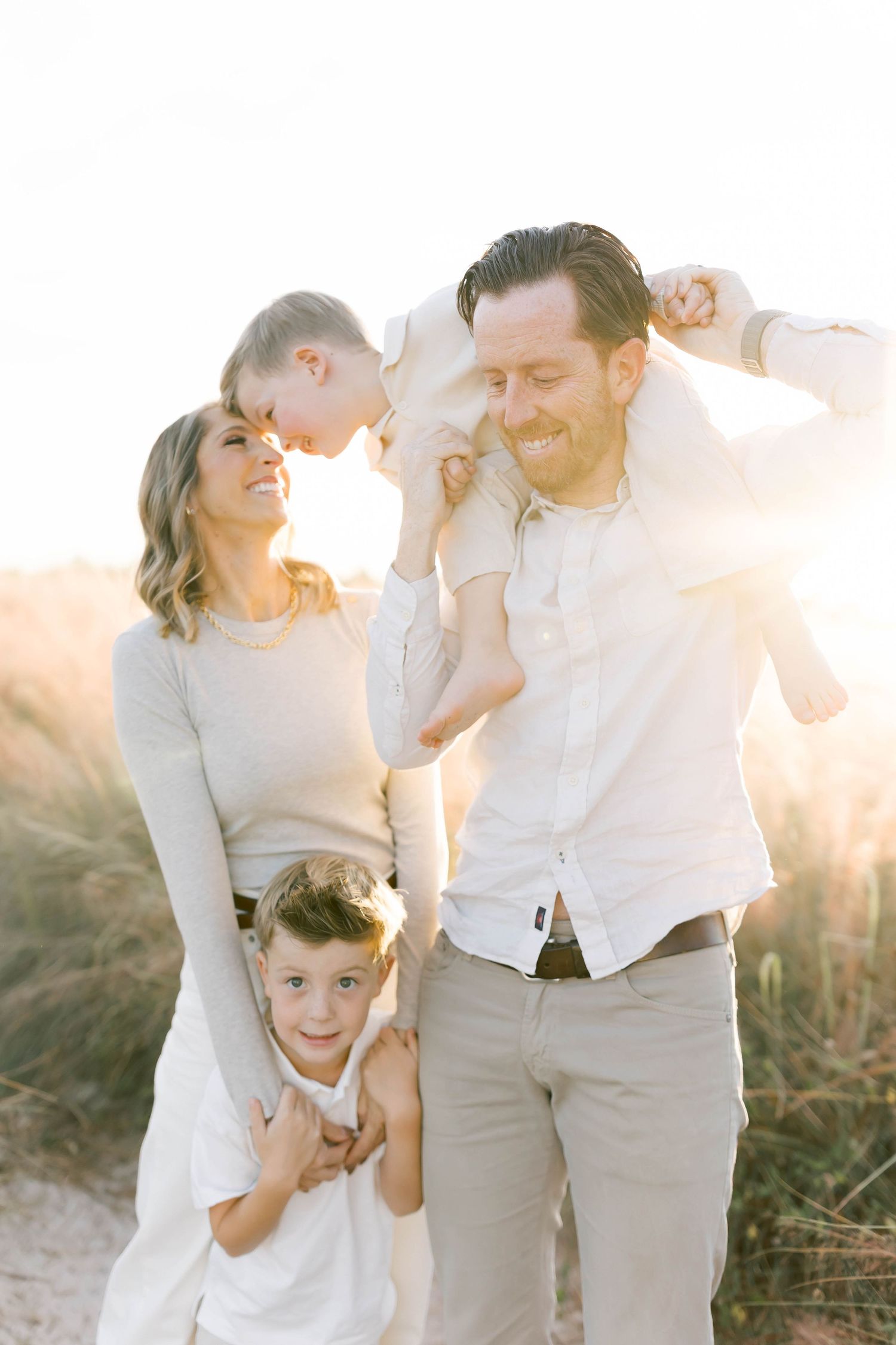 Family of four in white and neutral clothing share a candid moment in golden sunset light.