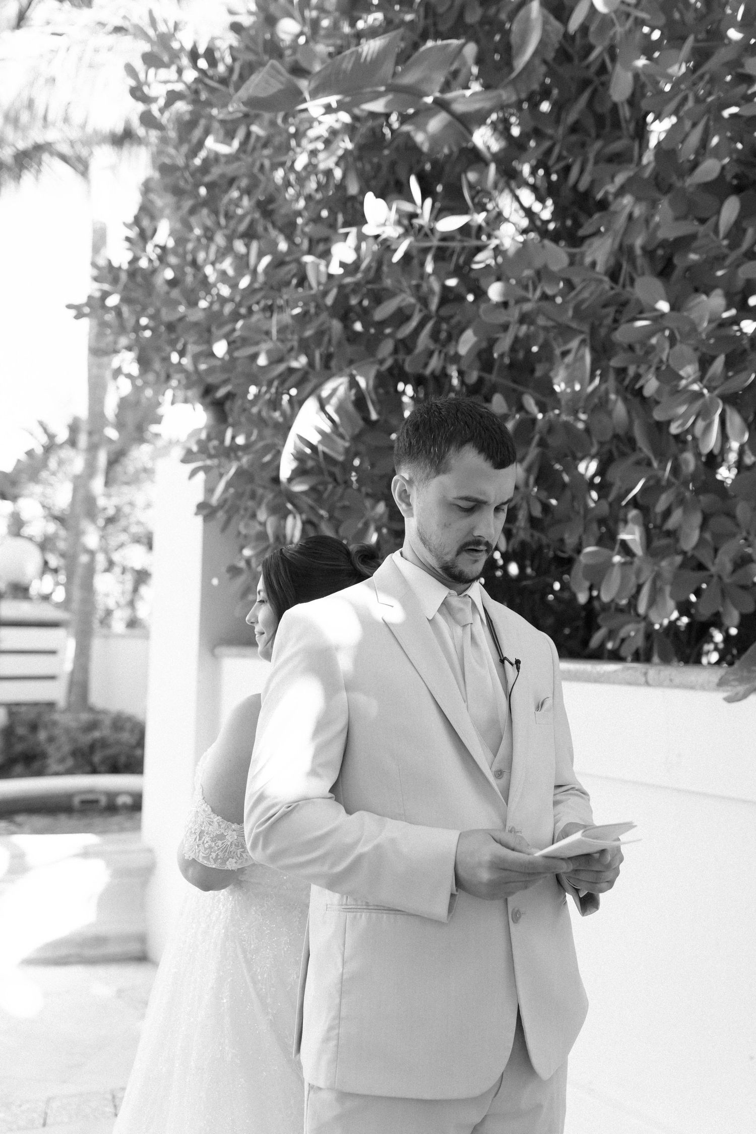 Black and white photo of groom in light suit reading vows outdoors near tropical greenery.