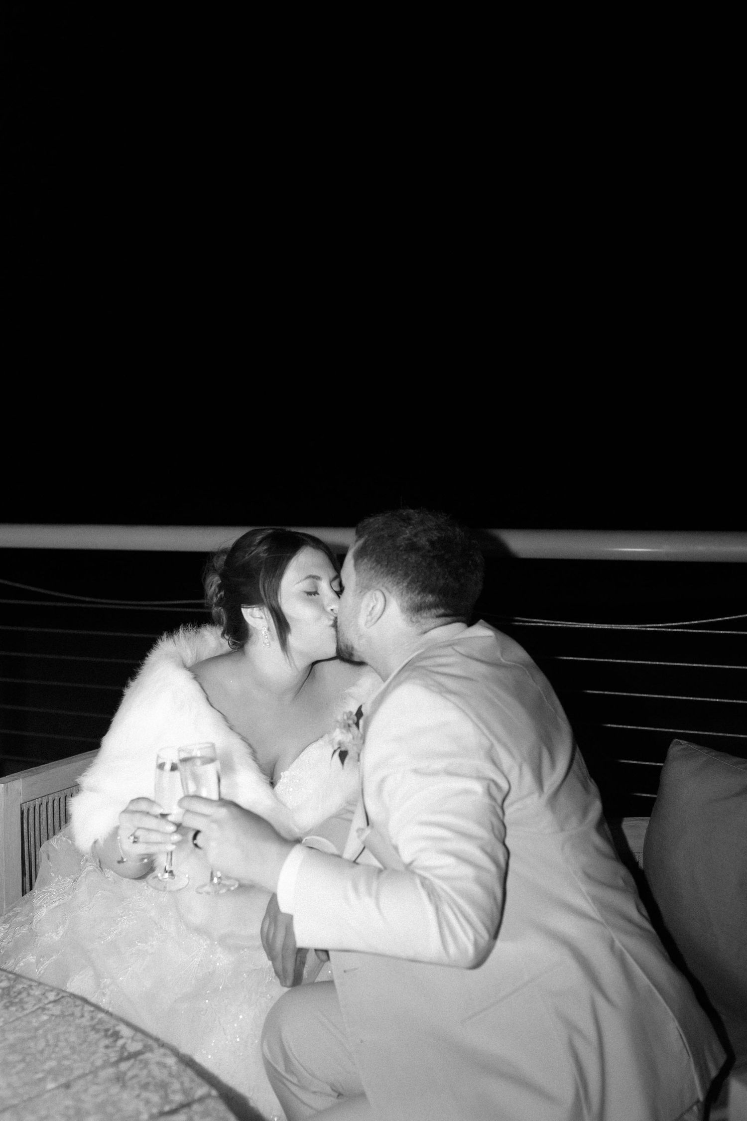 Black and white photo of bride and groom kissing while holding champagne glasses at night outdoors.