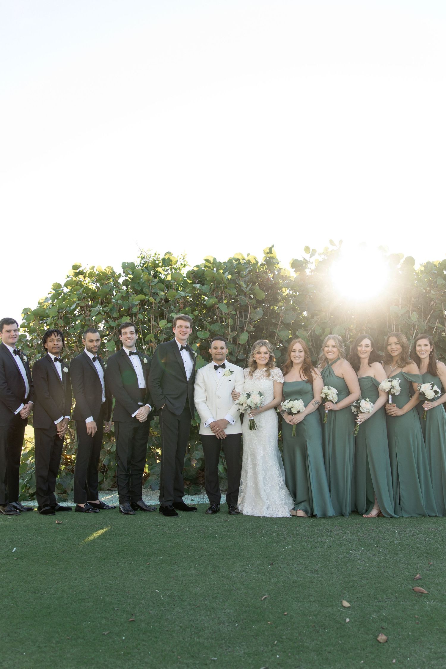 Wedding party in sage green dresses and black tuxedos lines up for formal photos outdoors.