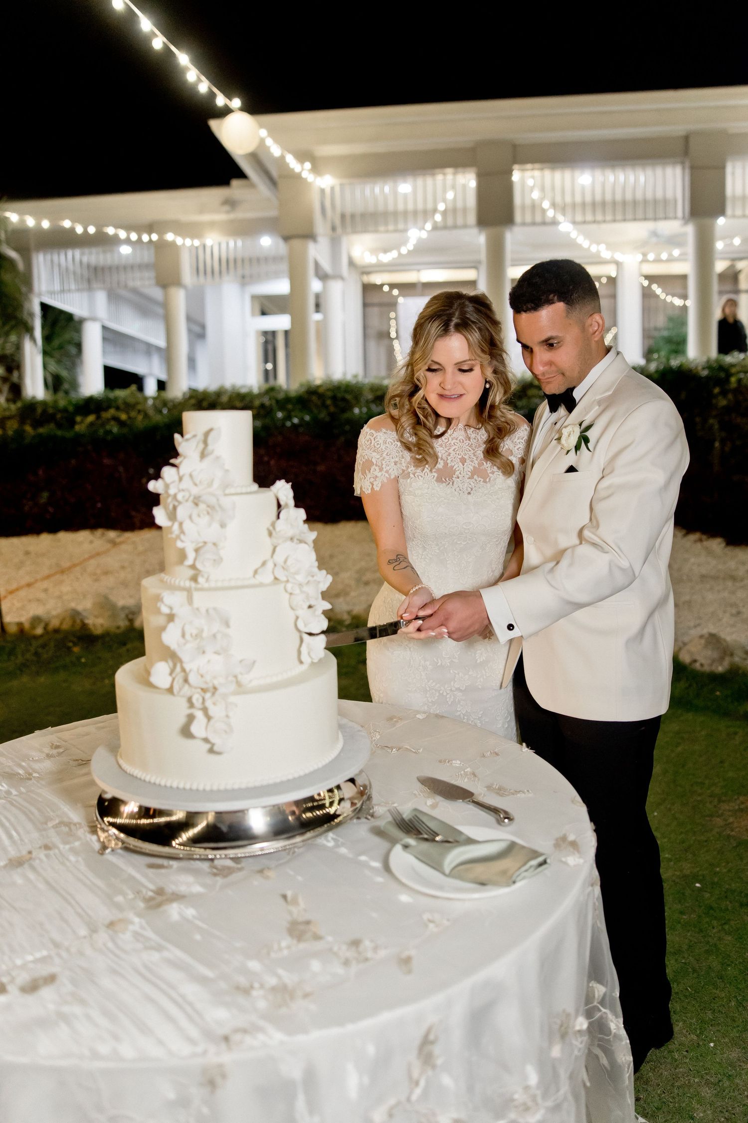 Couple cutting their elegant white wedding cake at an outdoor evening reception.