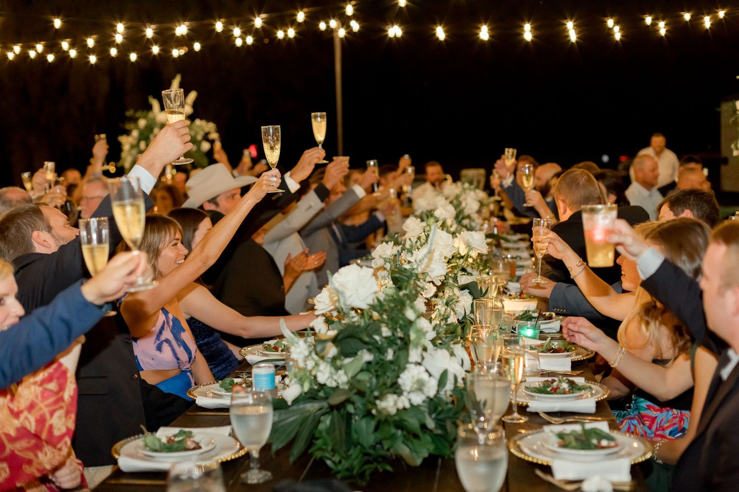 Wedding guests raising champagne glasses in a toast at an elegant outdoor reception with string lights and floral centerpieces.