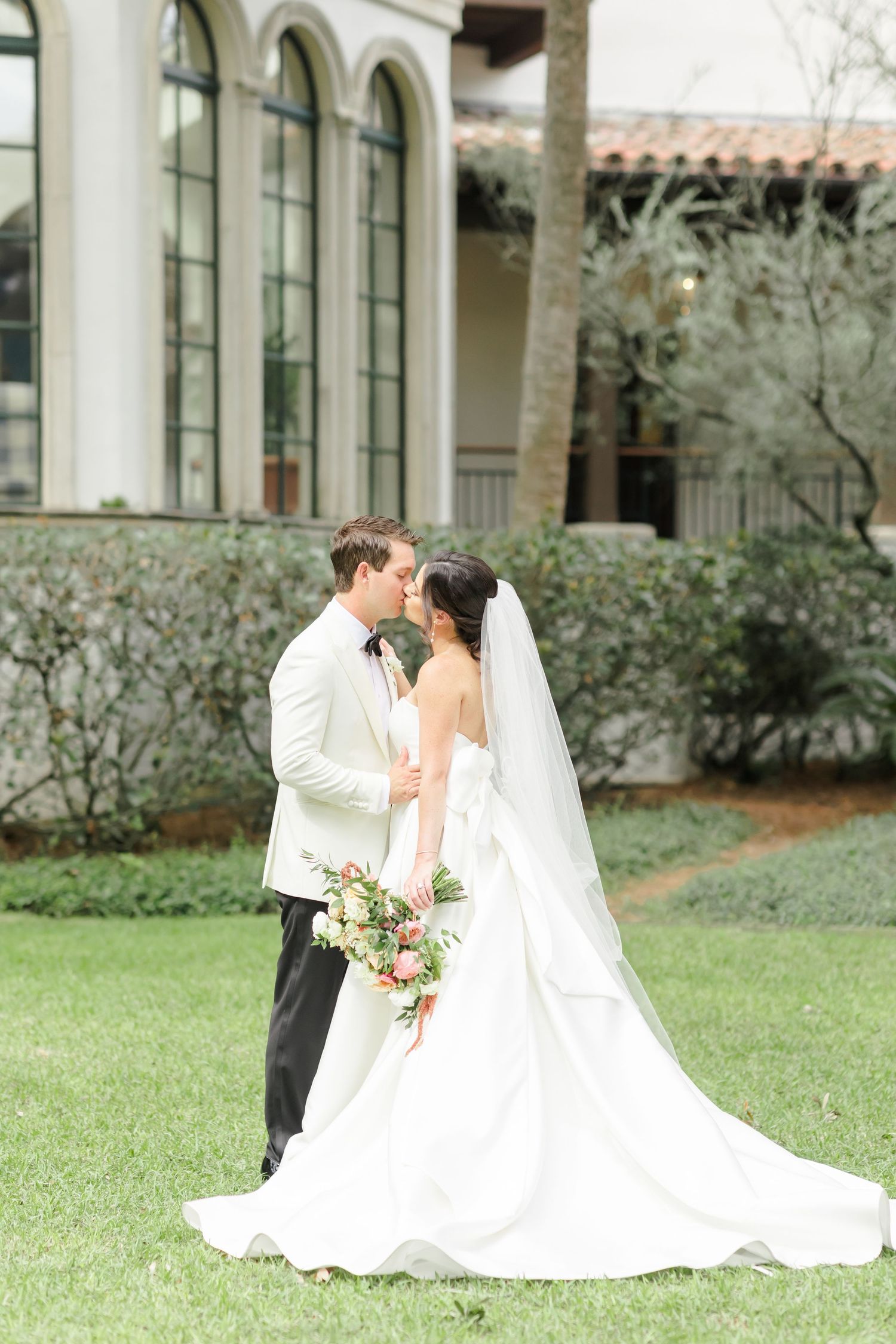 A romantic wedding couple shares intimate moments in a lush garden courtyard setting with white architecture.