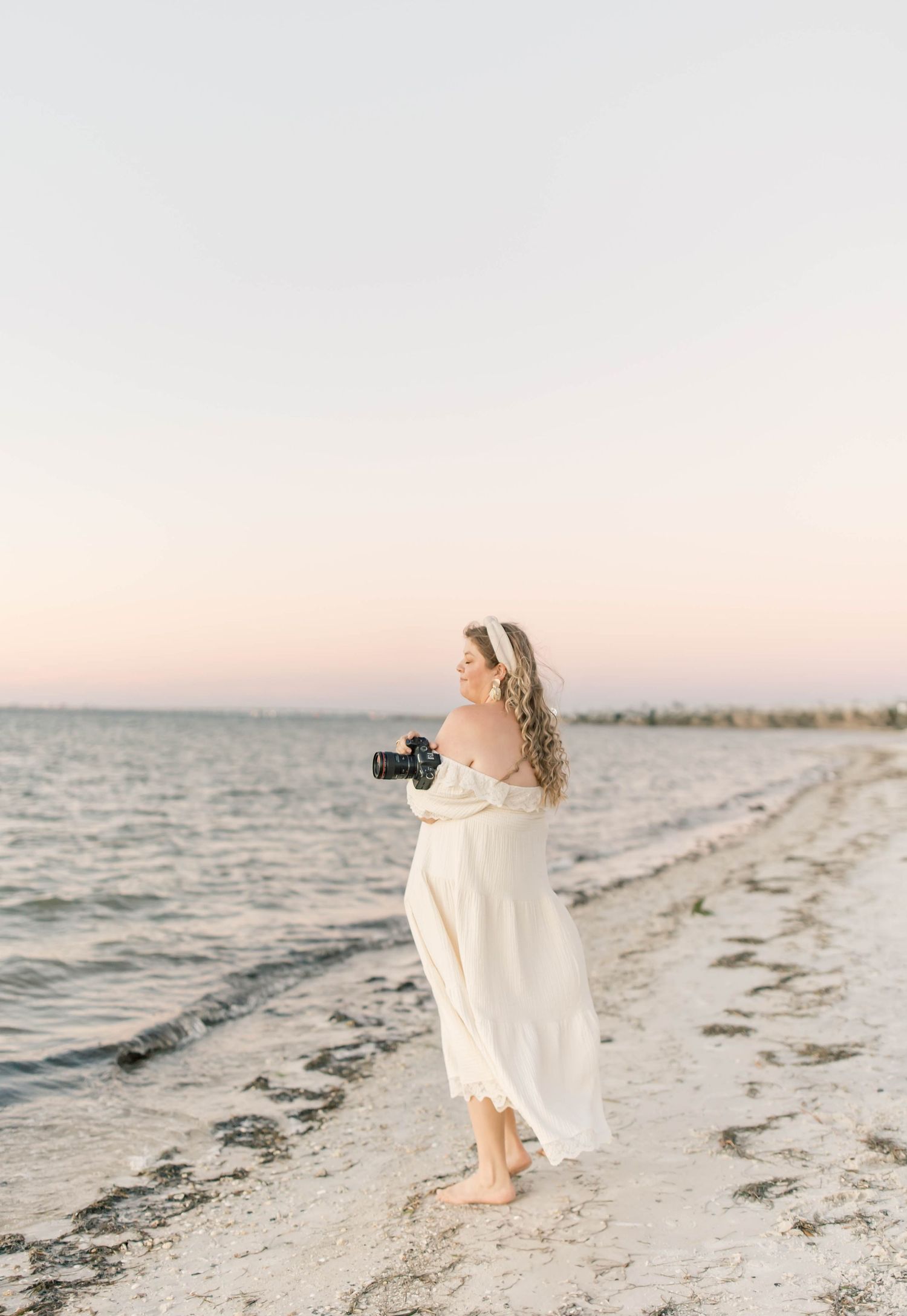 Woman in white dress holding camera on beach at sunset, waves gently lapping shore behind her.