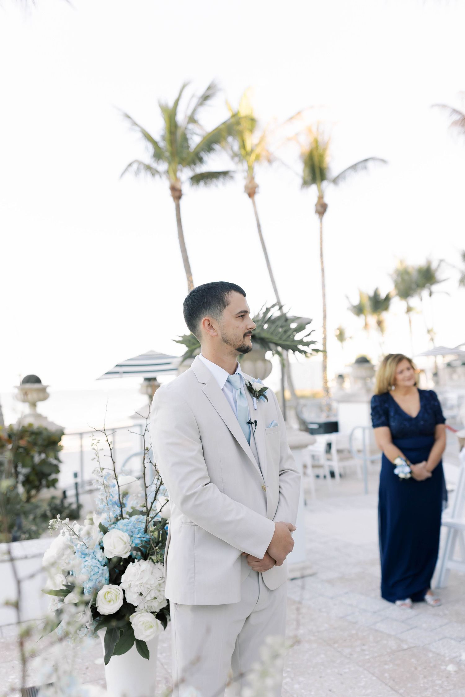 Groom in beige suit stands at outdoor altar with blue and white floral arrangements, palm trees and ocean in background.
