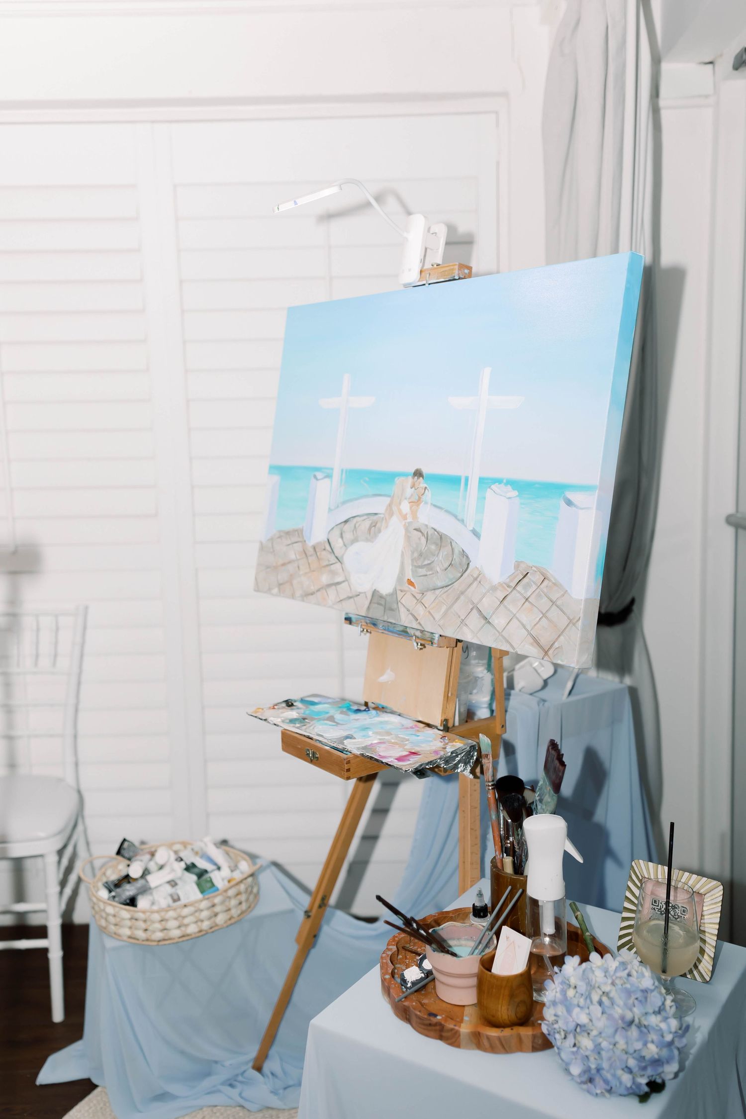 A wedding reception easel displaying a blue ocean painting with art supplies on a draped table nearby.