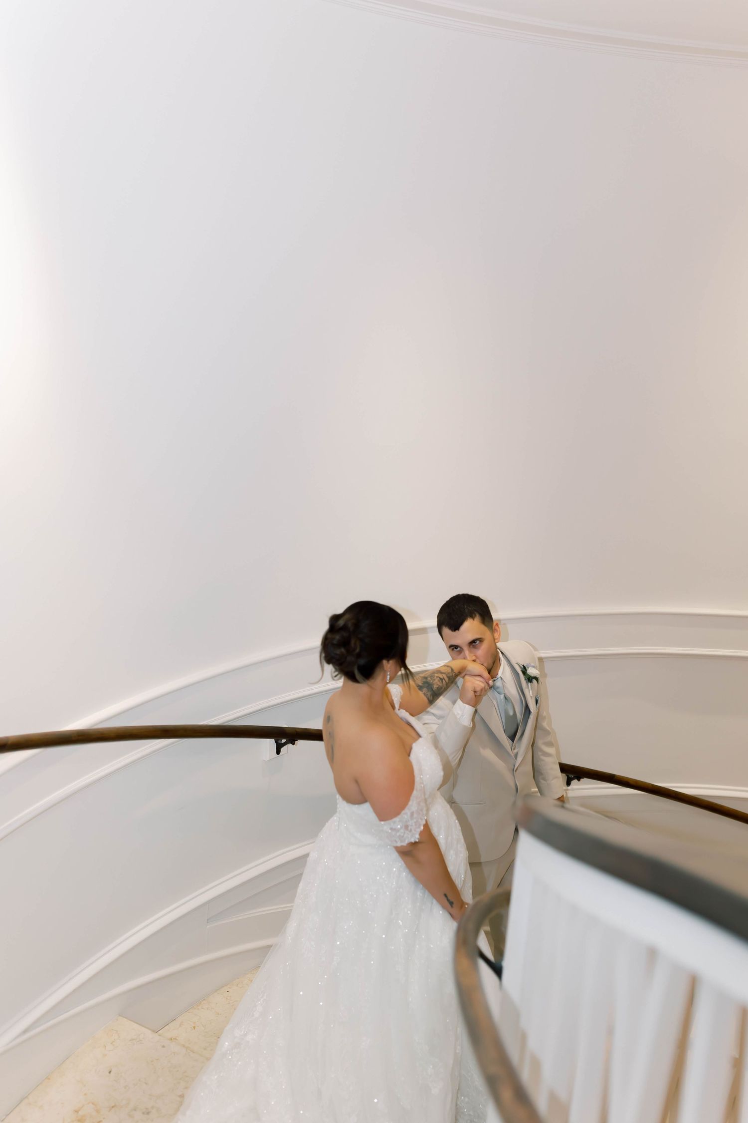 Groom kisses bride's hand on elegant curved staircase with white walls in a bright indoor venue.