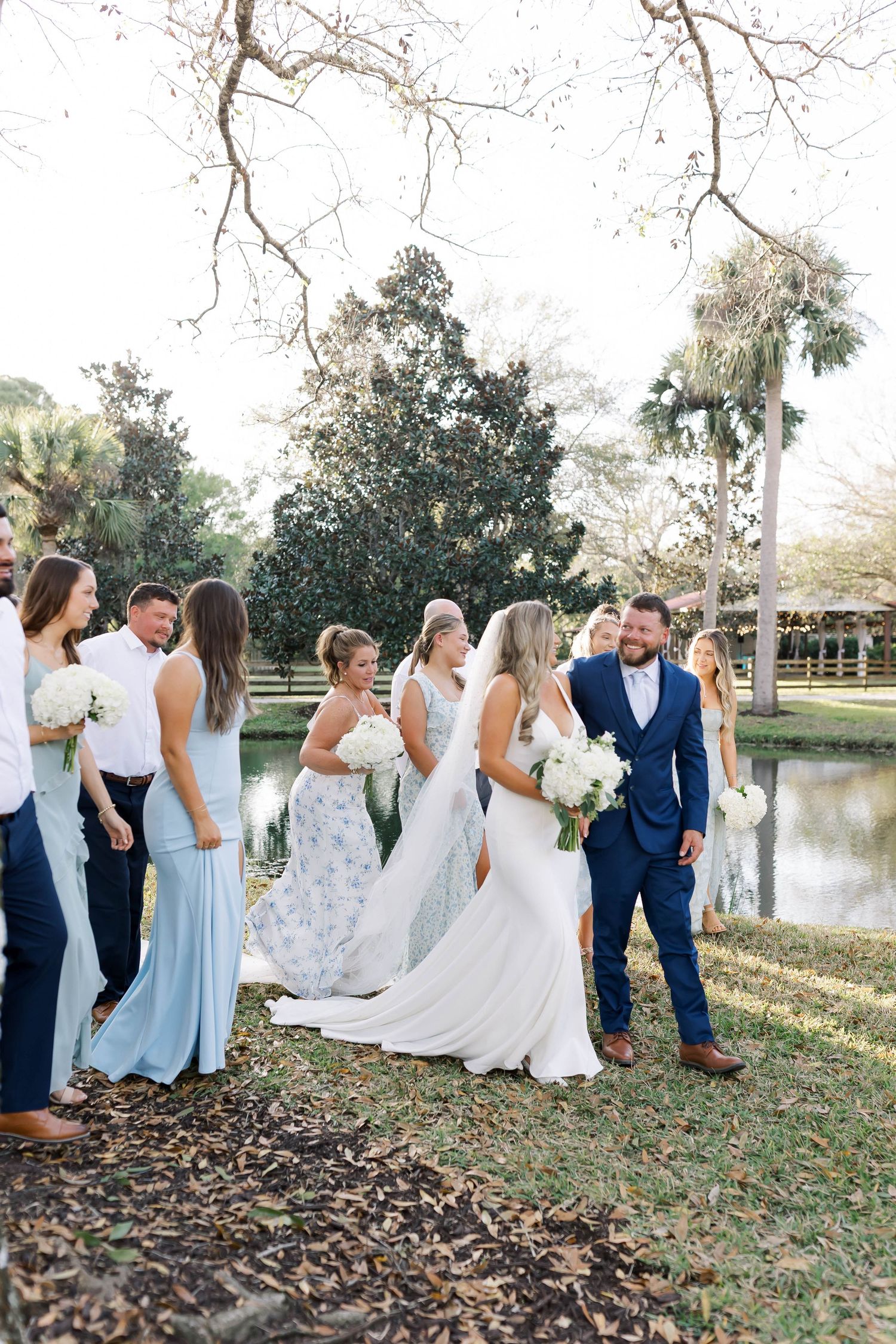 Wedding group poses by lakeside with trees in background wearing coordinated attire.
