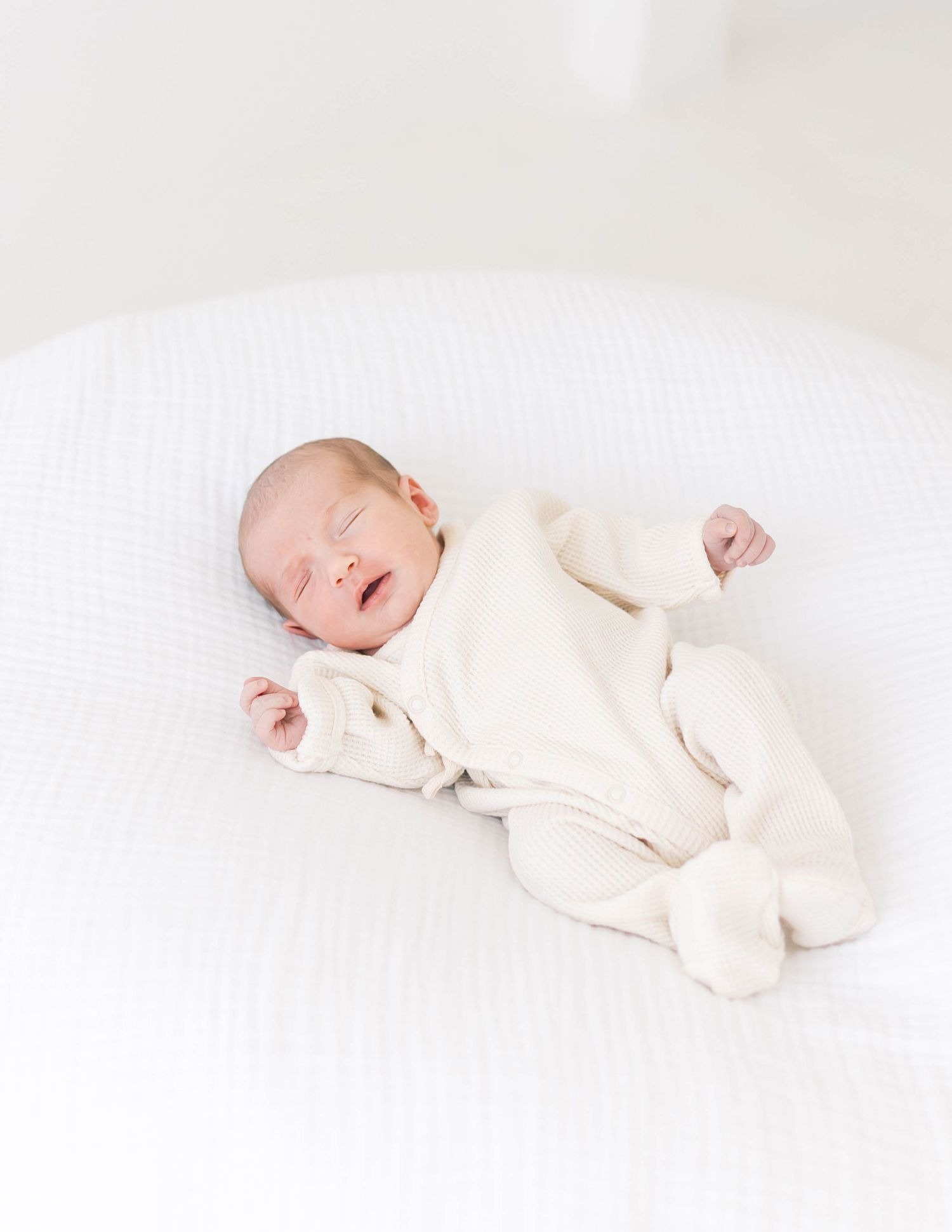 Sleeping newborn baby in cream colored outfit lying on white backdrop in minimalist photography series.