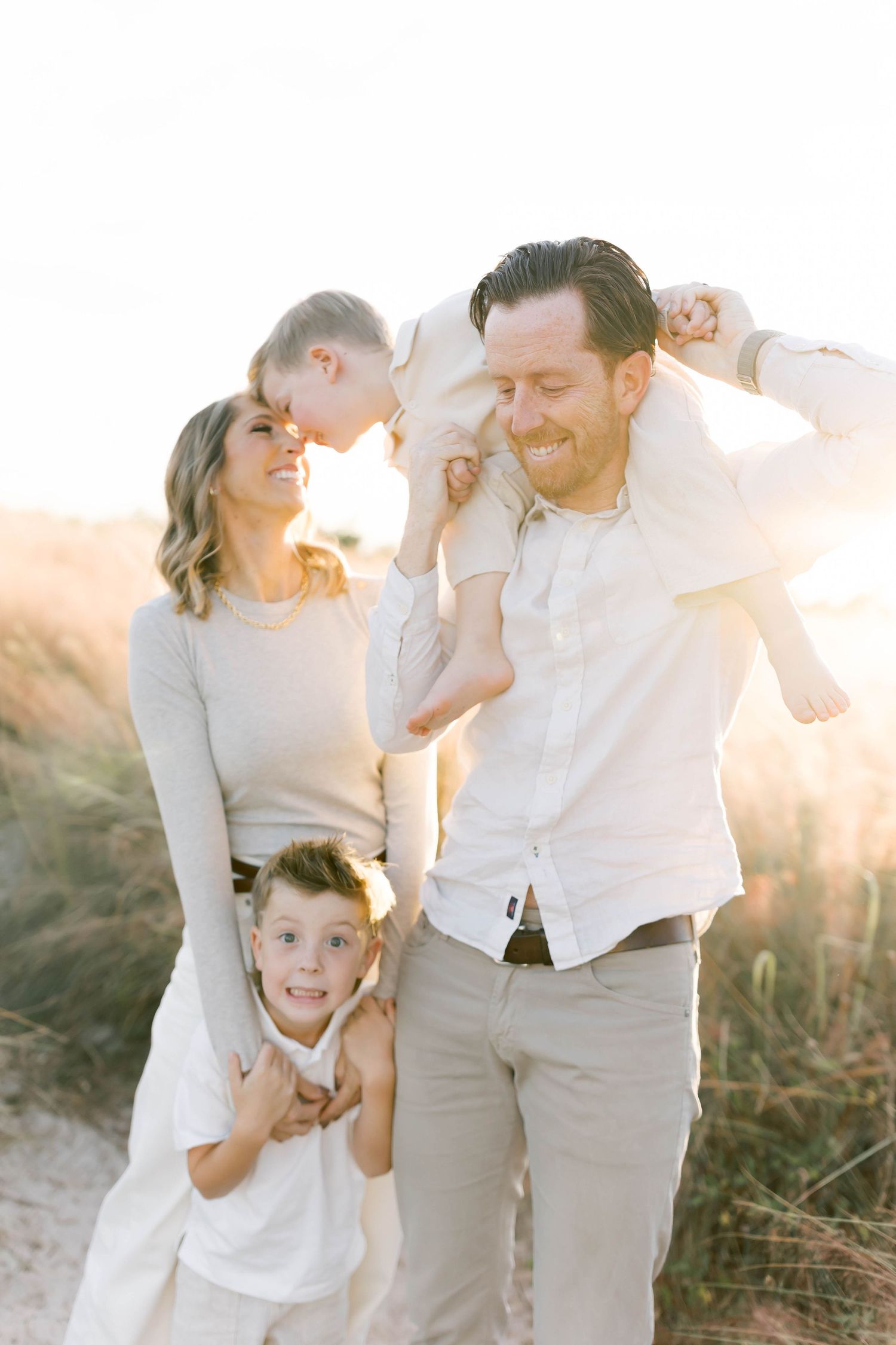 A family in coordinated white and beige outfits plays together during golden hour in a field.
