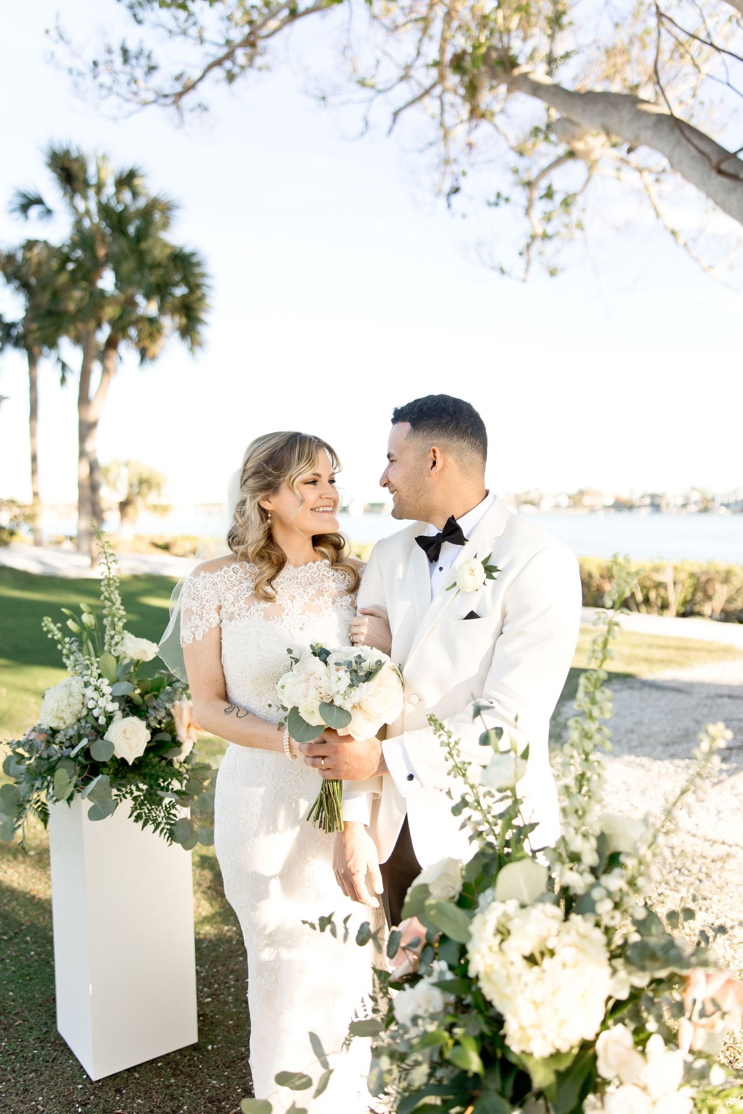 Elegant wedding couple poses outdoors with white floral arrangements and greenery.
