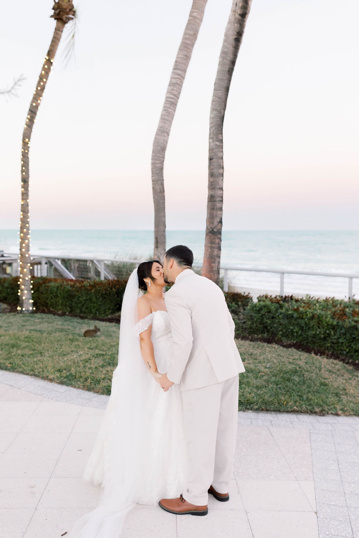 Bride and groom share a romantic kiss by the ocean at sunset, surrounded by palm trees at a beachside wedding.