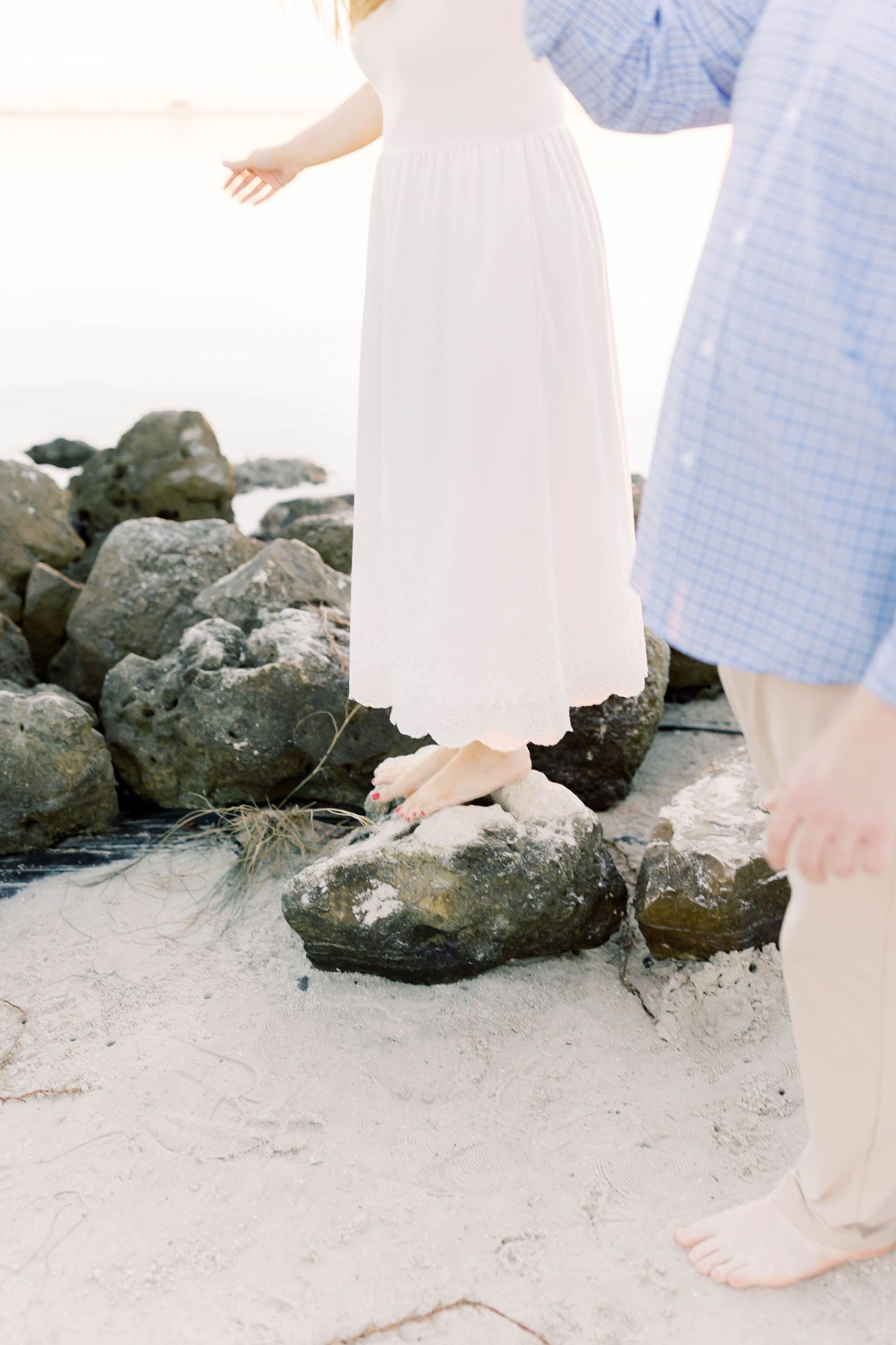 Barefoot couple stands together on sandy beach with rocky shoreline.