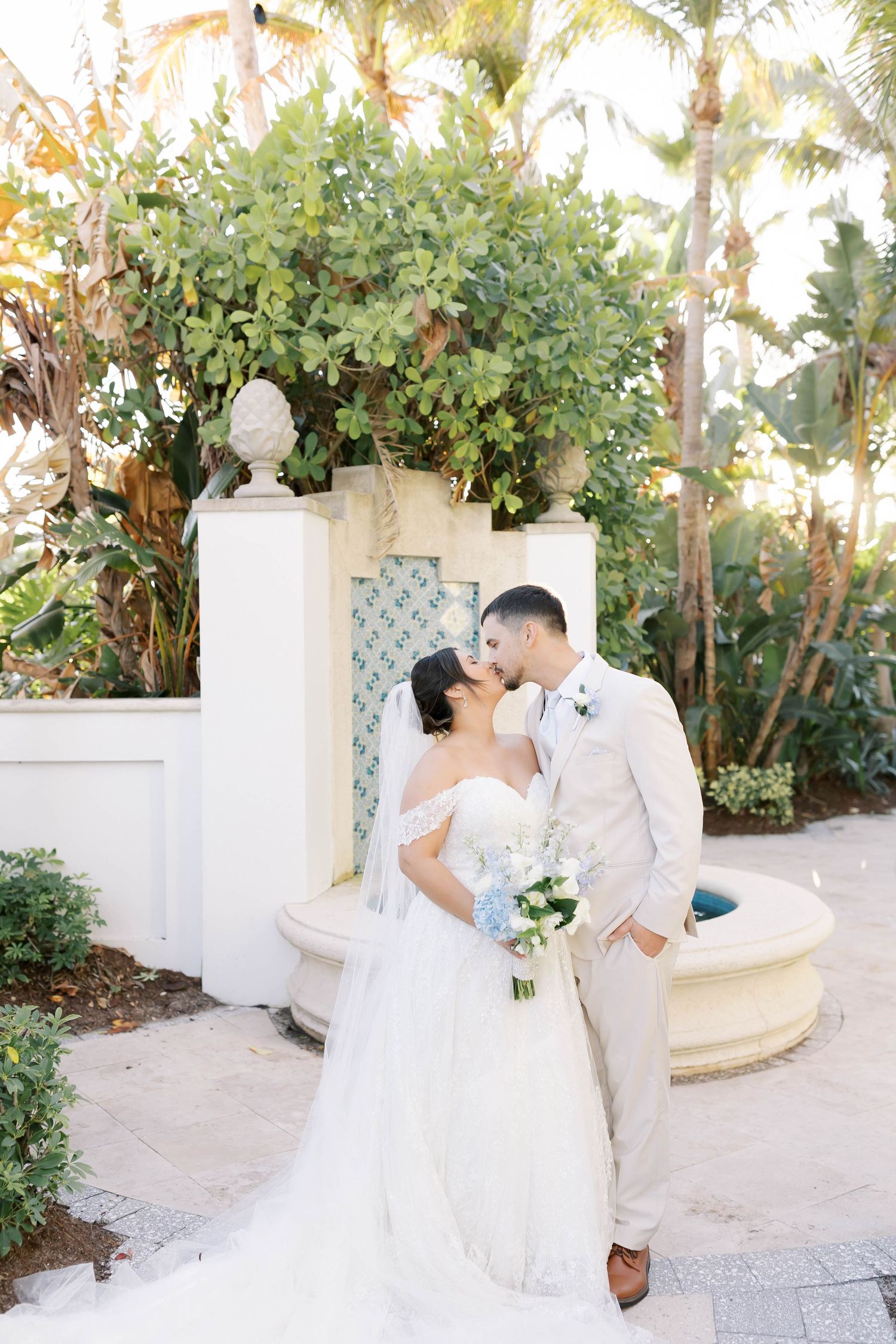 Newlyweds share a romantic kiss in a lush tropical garden setting, bride holding a blue and white bouquet.