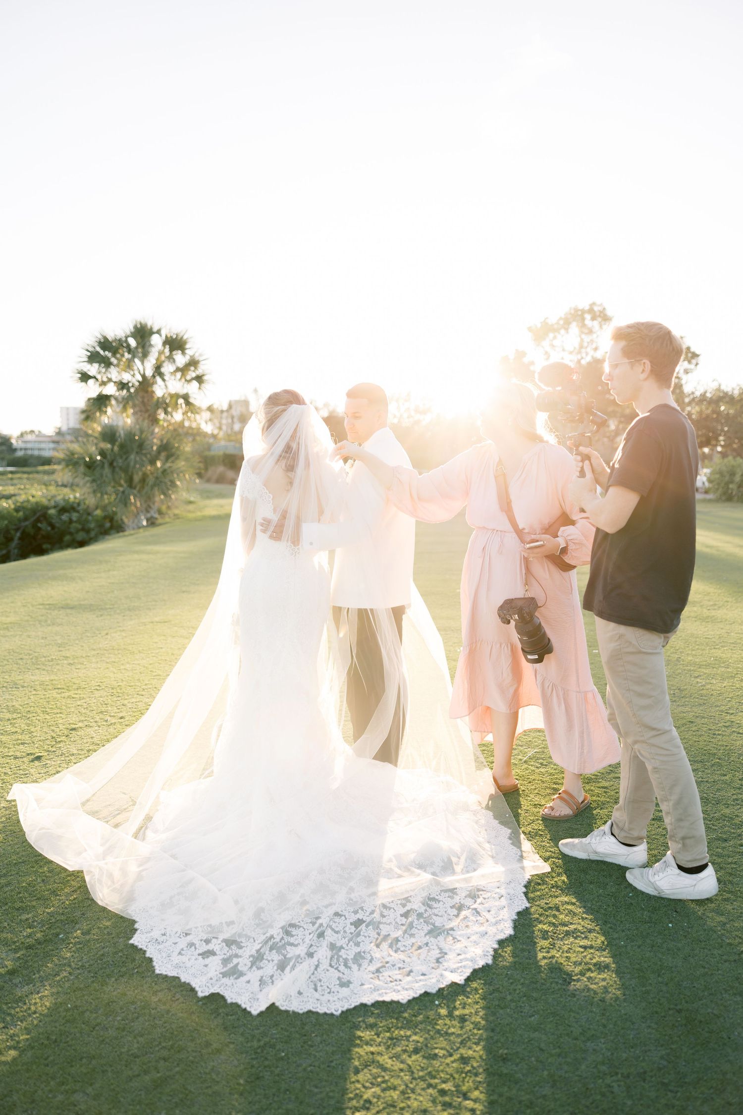 Bride in white gown with lace veil poses with groom and wedding photographers on green lawn at sunset.