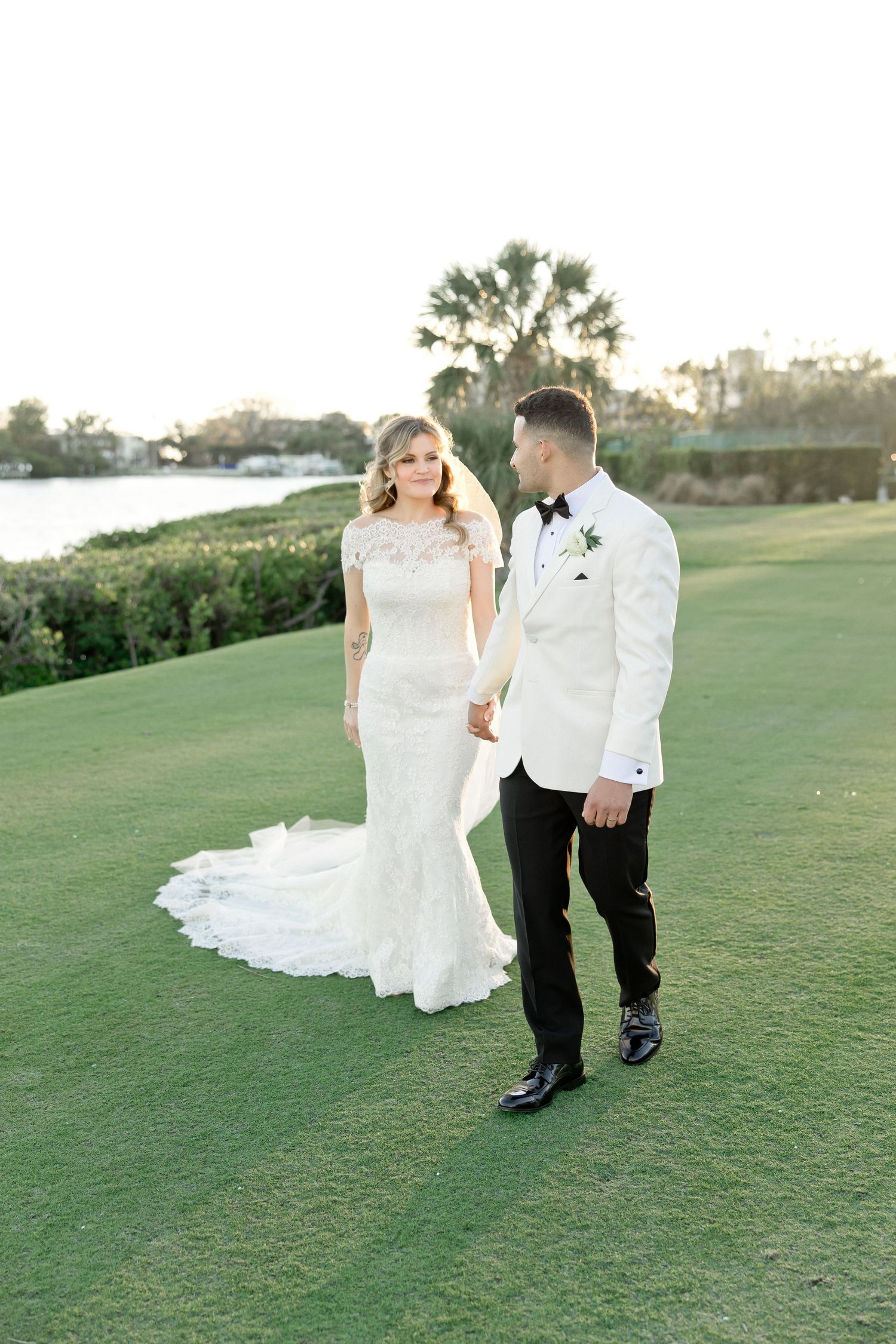 Bride in lace mermaid gown and groom in white tuxedo hold hands on a lush green golf course at sunset.