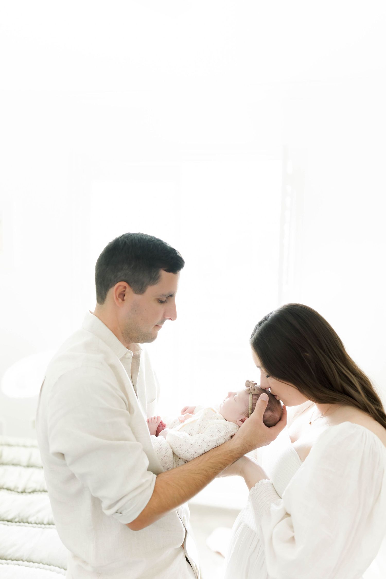 Parents in white clothing tenderly hold their newborn baby against bright window.