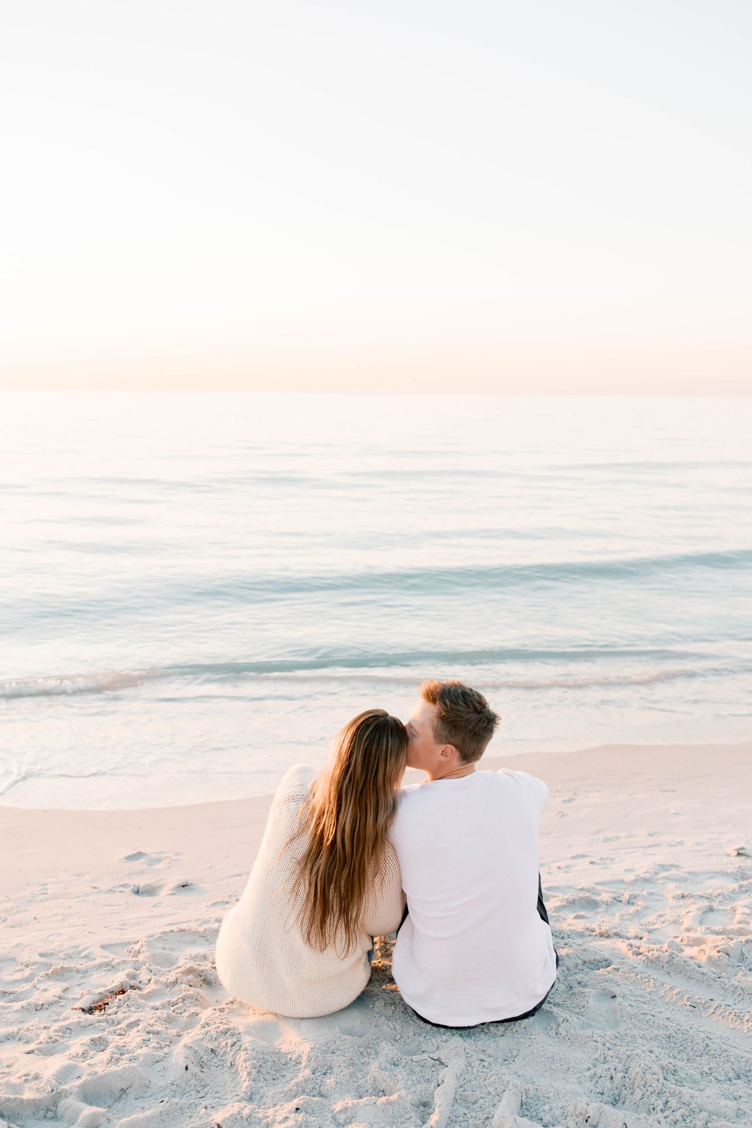 A couple sits together on a white sandy beach at sunset sharing an intimate moment.