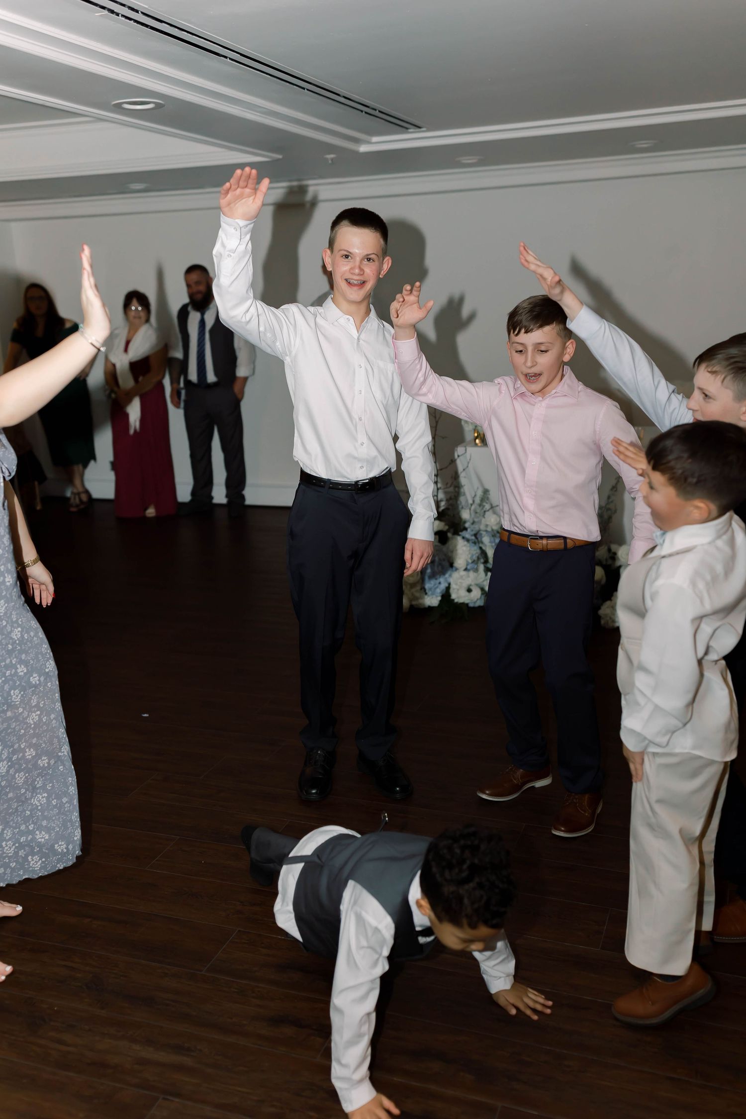 Energetic children celebrate on the wedding reception dance floor with arms raised in a bright white venue.