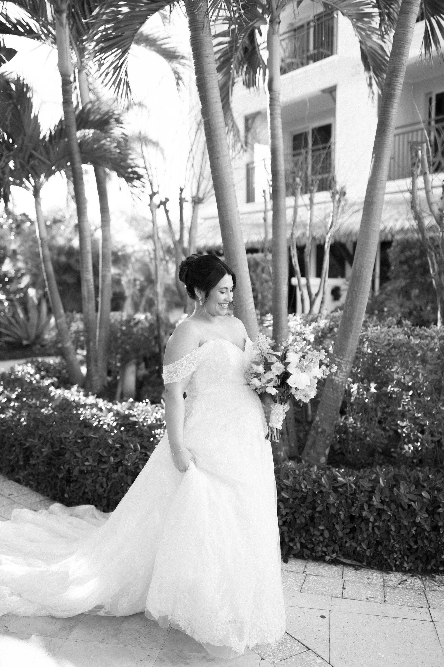 Black and white portrait of bride in off-shoulder gown holding bouquet among palm trees.