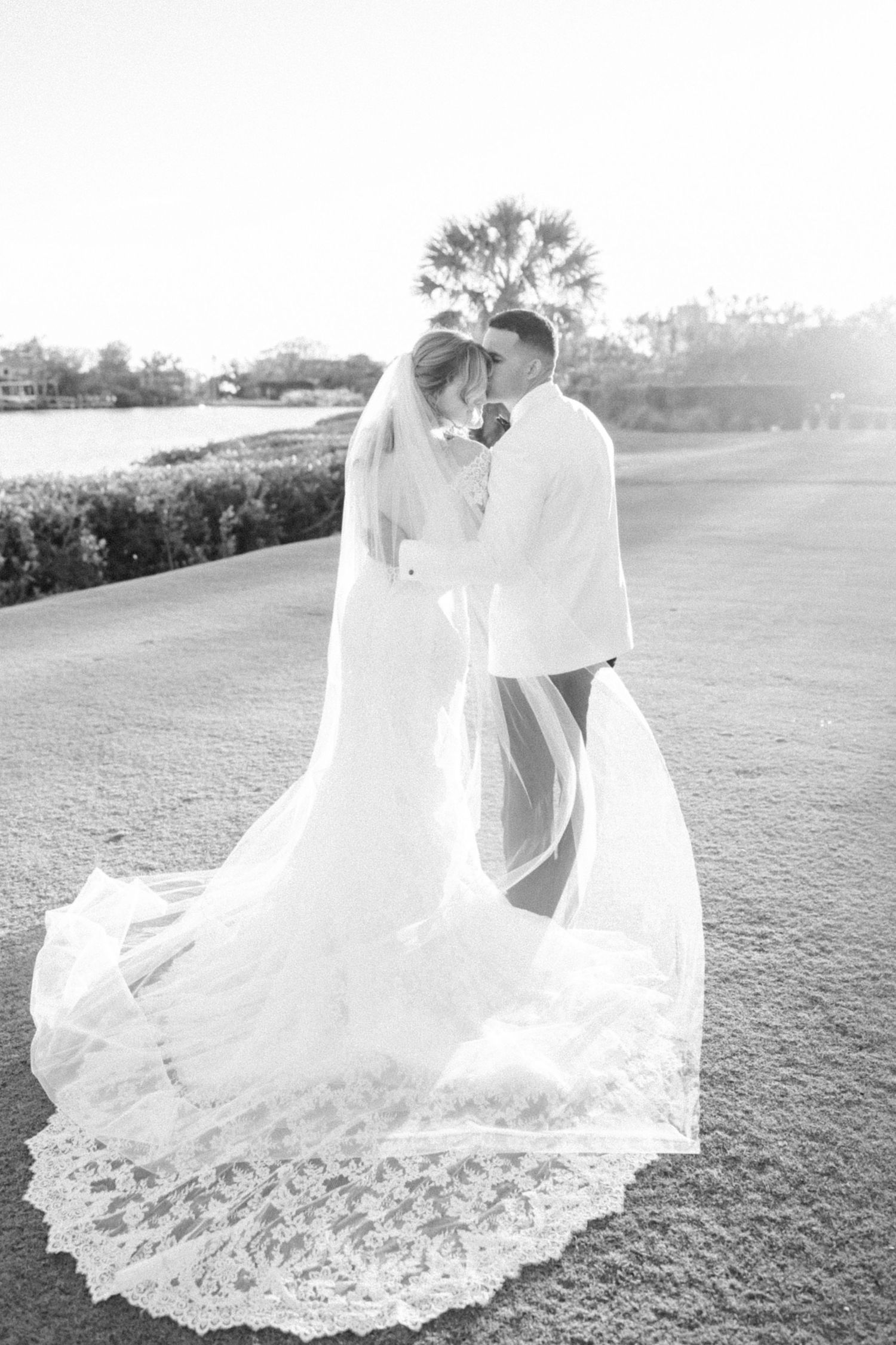 Flowing lace wedding dress train spread across green grass during sunset photoshoot.