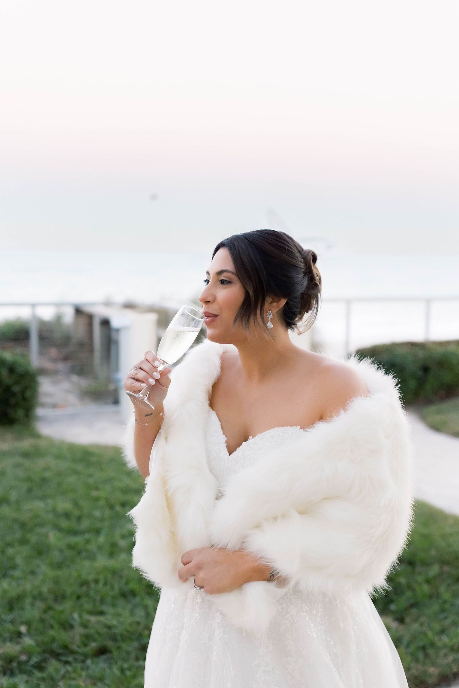 Elegant bride in white gown and faux fur wrap sips champagne near the ocean at sunset.