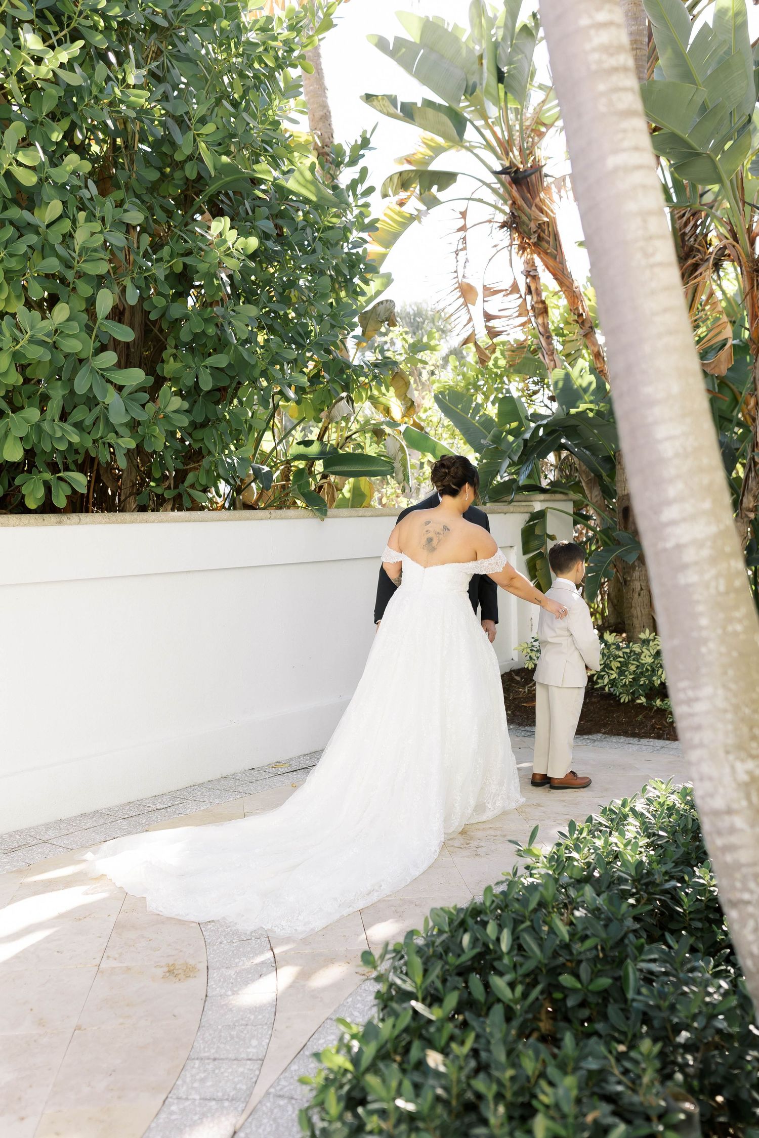 Bride in off-shoulder gown with long train walks with young boy in garden setting at tropical wedding venue.
