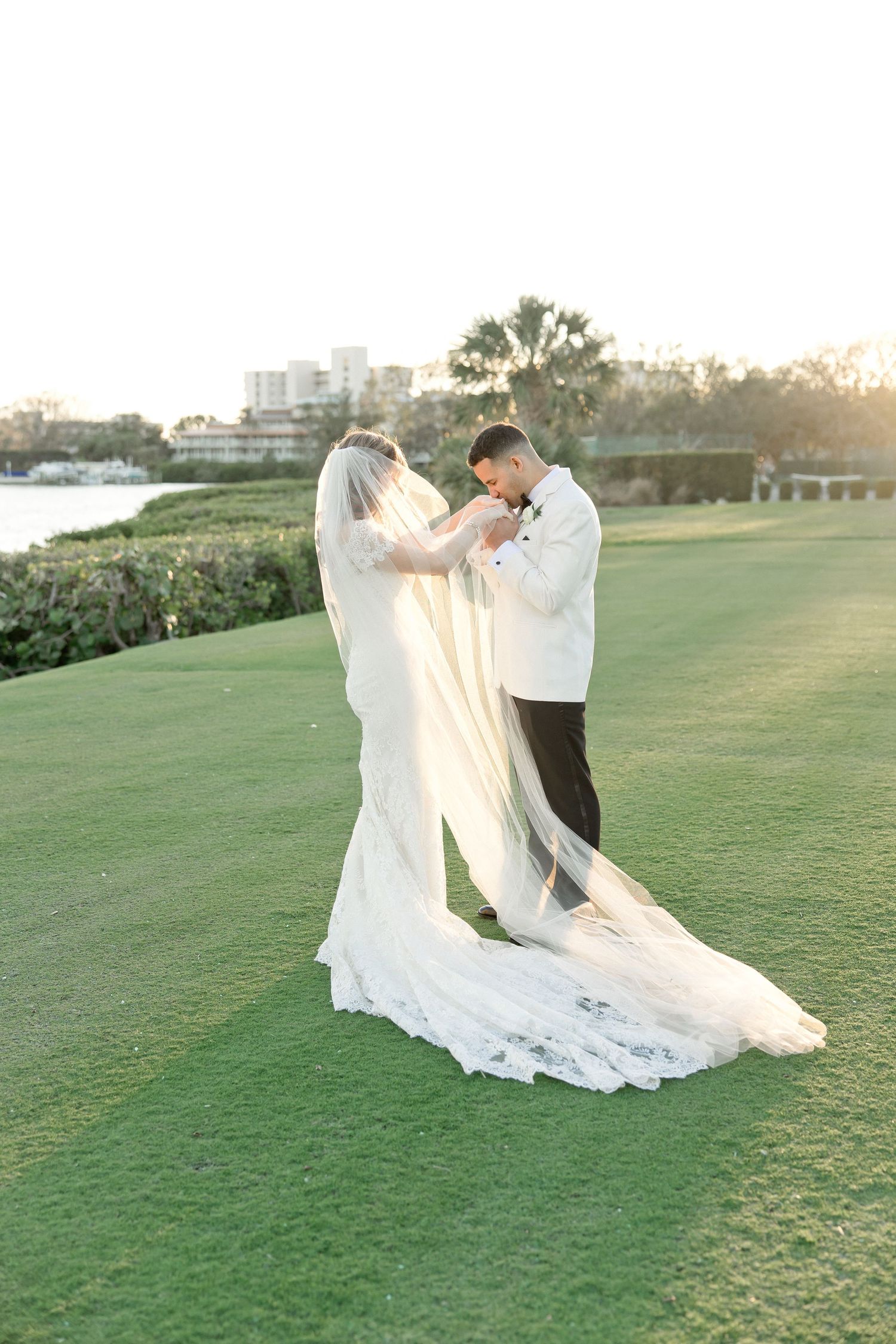 A couple in wedding attire dances gracefully on a green lawn at sunset with a flowing white veil creating elegant movement.