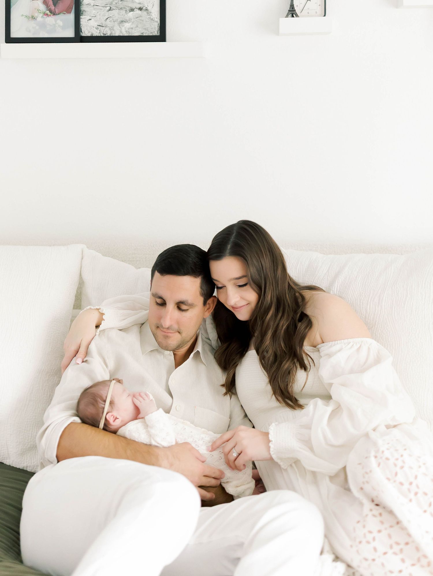 Family cuddles together on white couch with newborn baby in bright room.