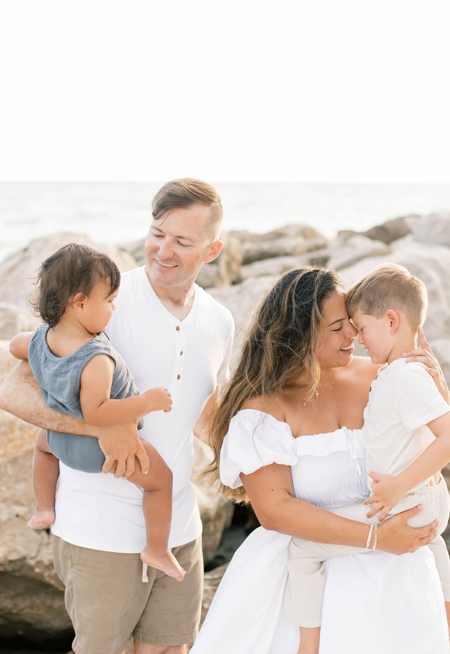 Family wearing white and neutral colors embracing and laughing together on a beach.