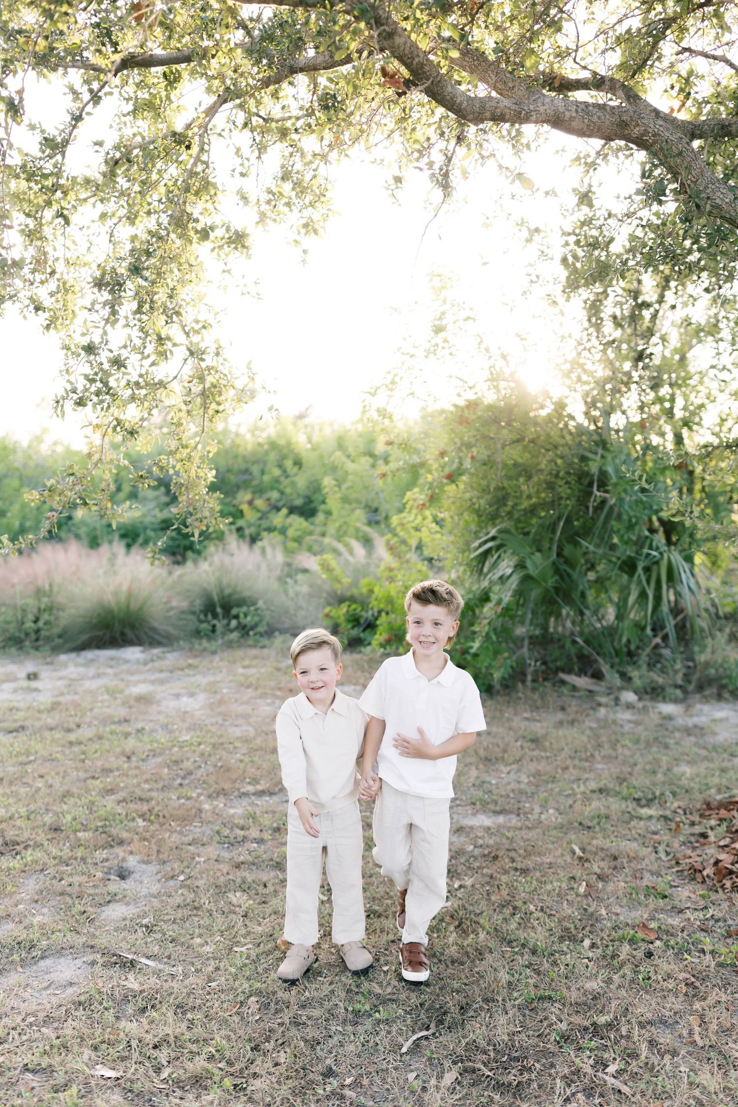 Two children in matching white outfits stand together under a large tree branch in a natural setting.