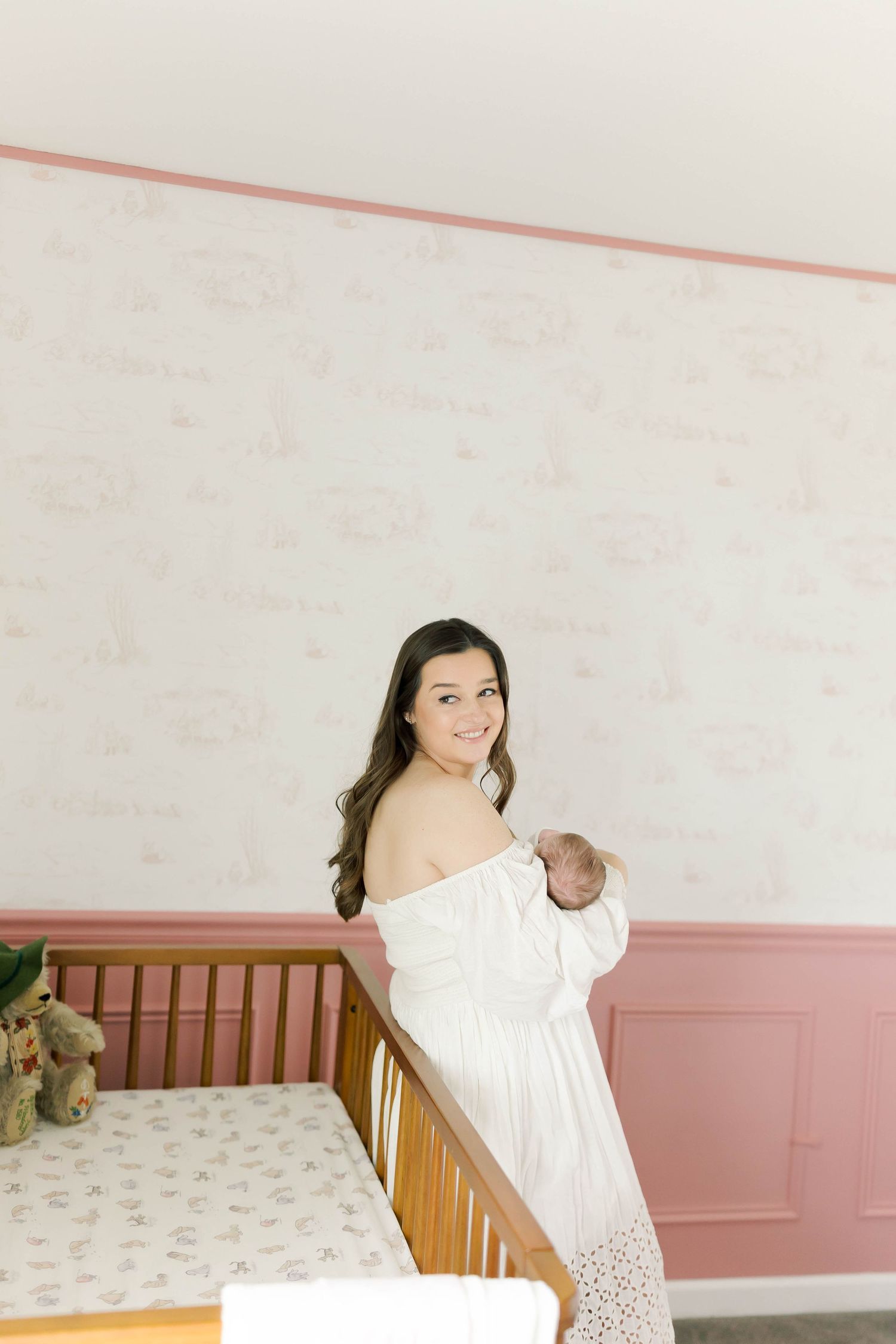 Someone in a white lace dress holds a baby next to a wooden crib in a pink and white nursery.