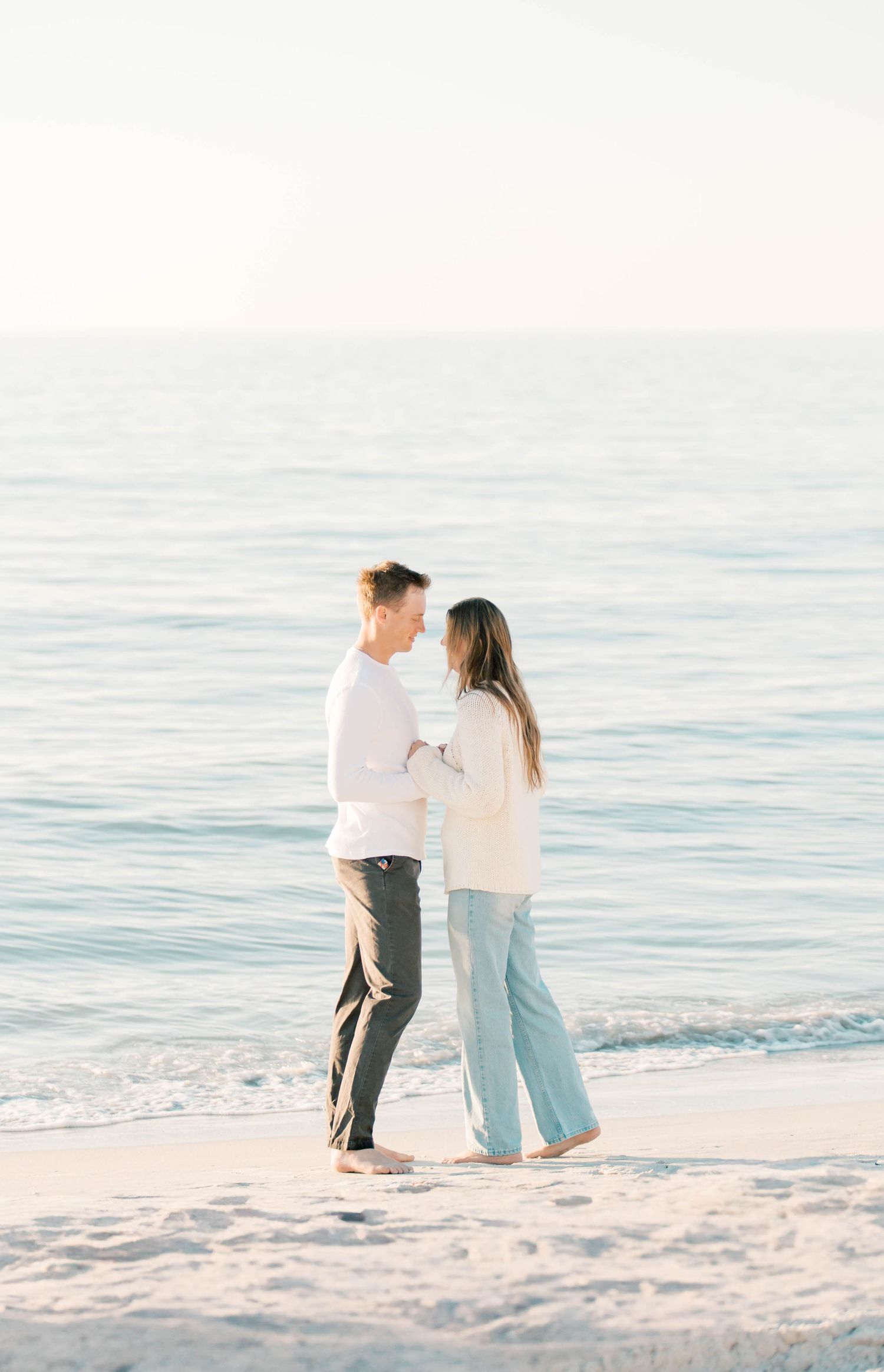 A couple stands close together on a beach with gentle waves and soft sunset lighting.